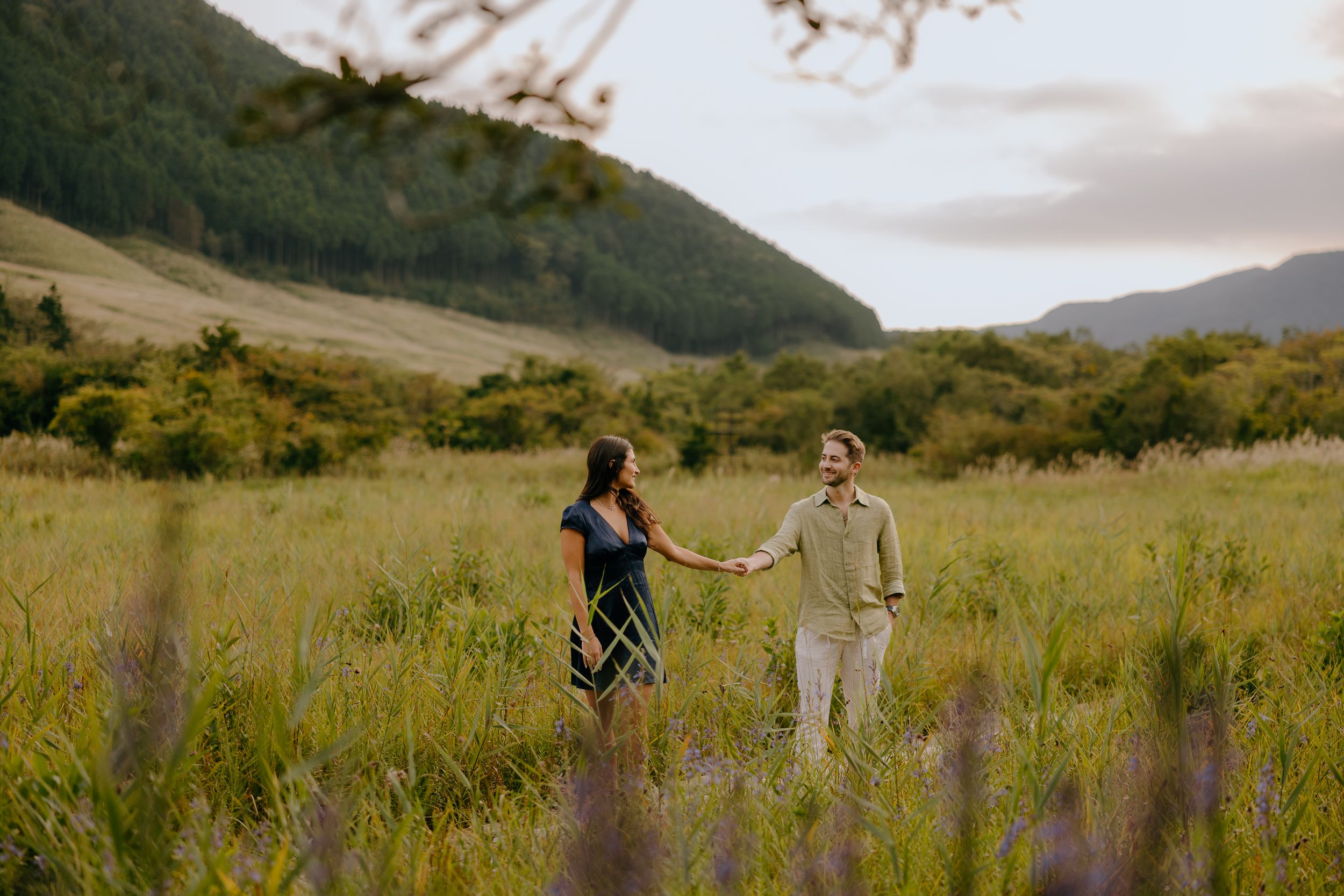 Couple holding hands while walking through scenic mountain meadow with golden grass and wildflowers at sunset.