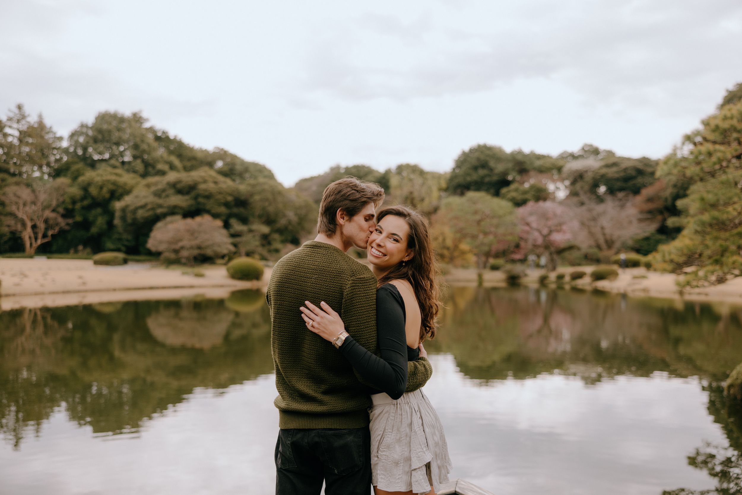 A loving embrace between two people by a serene pond surrounded by trees on an autumn day.