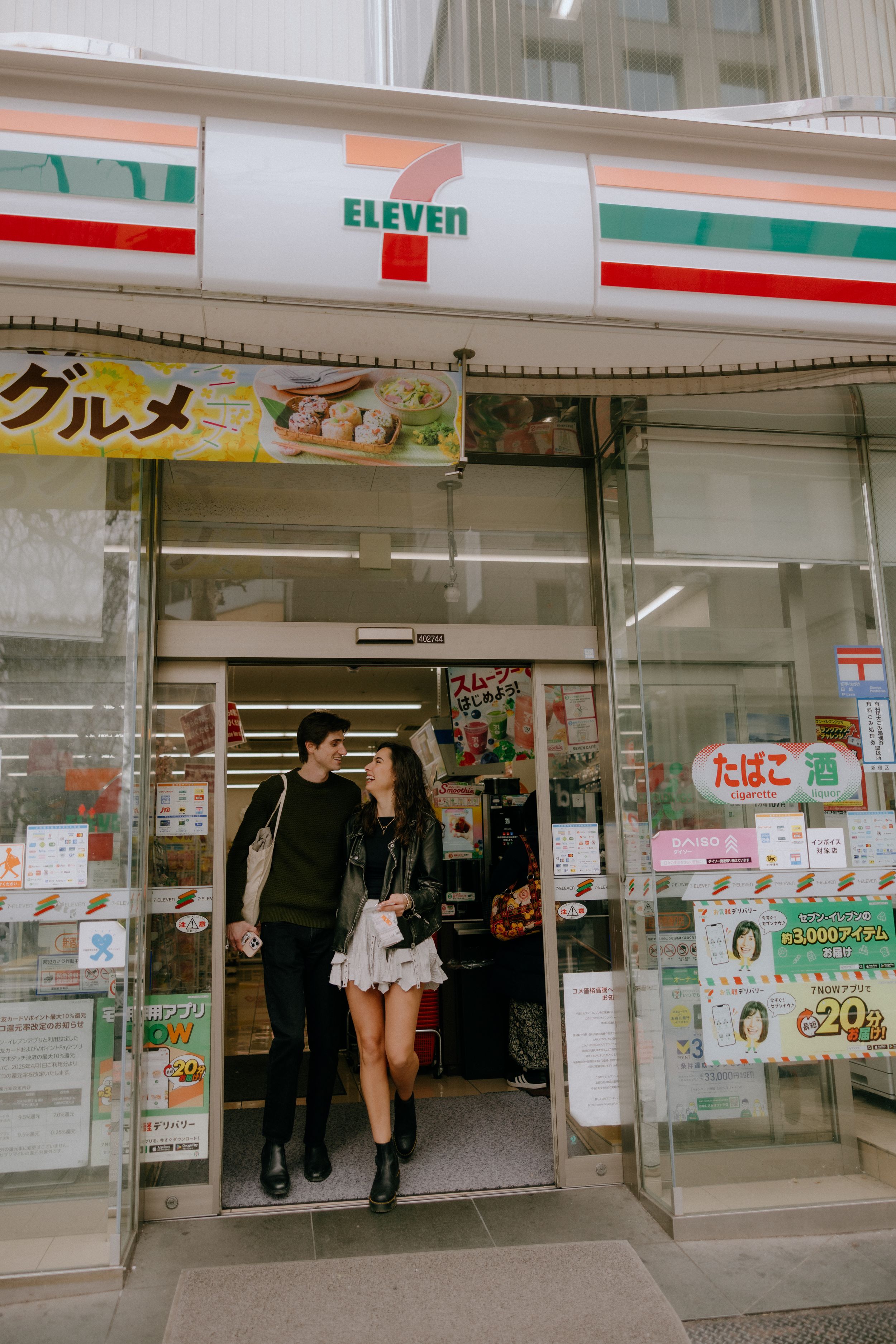 Customers exit a 7-Eleven convenience store in Japan with shopping bags and promotional signage in the storefront.