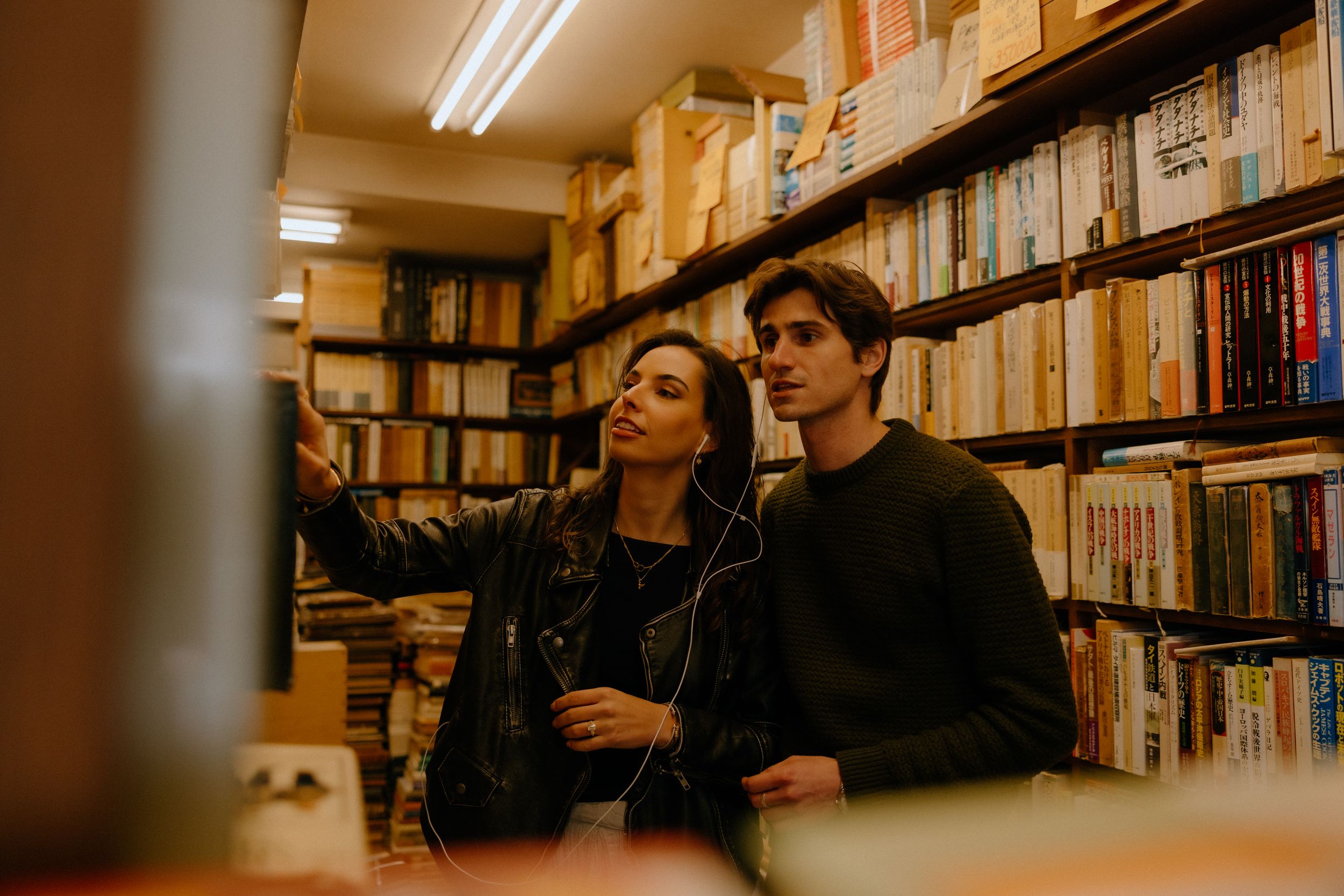 Two friends share earbuds while browsing books in a dimly lit library or bookstore.