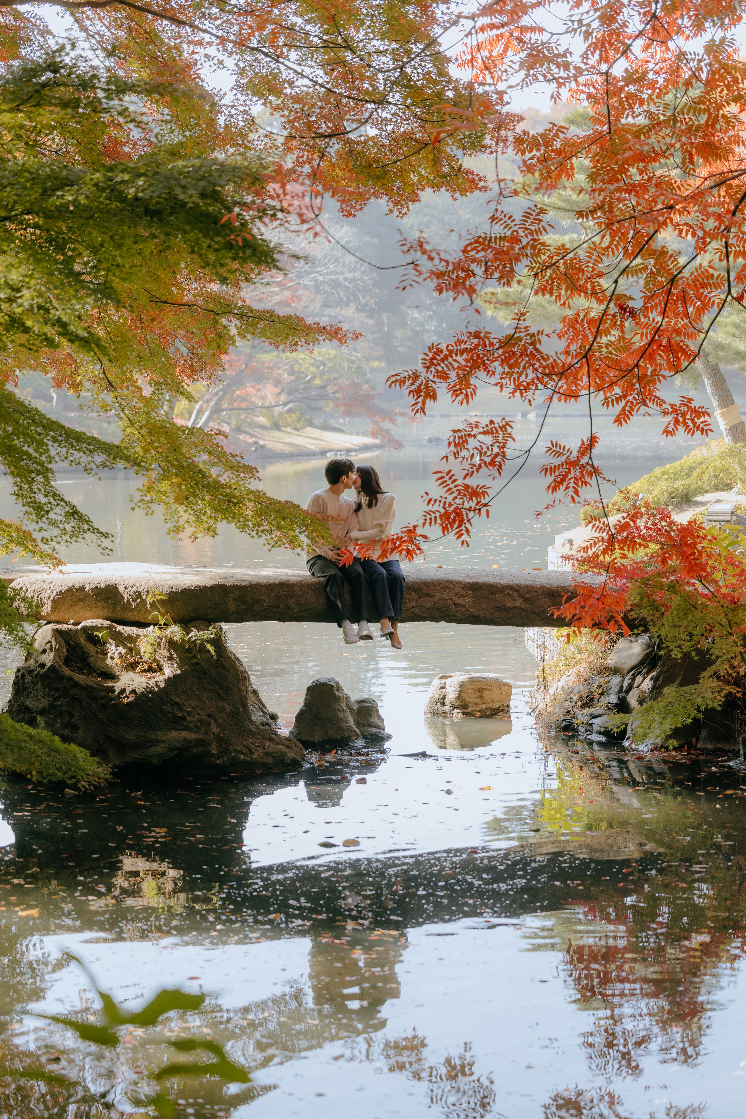 Silhouettes sit on a curved tree branch over a tranquil pond surrounded by autumn foliage with orange and green leaves.