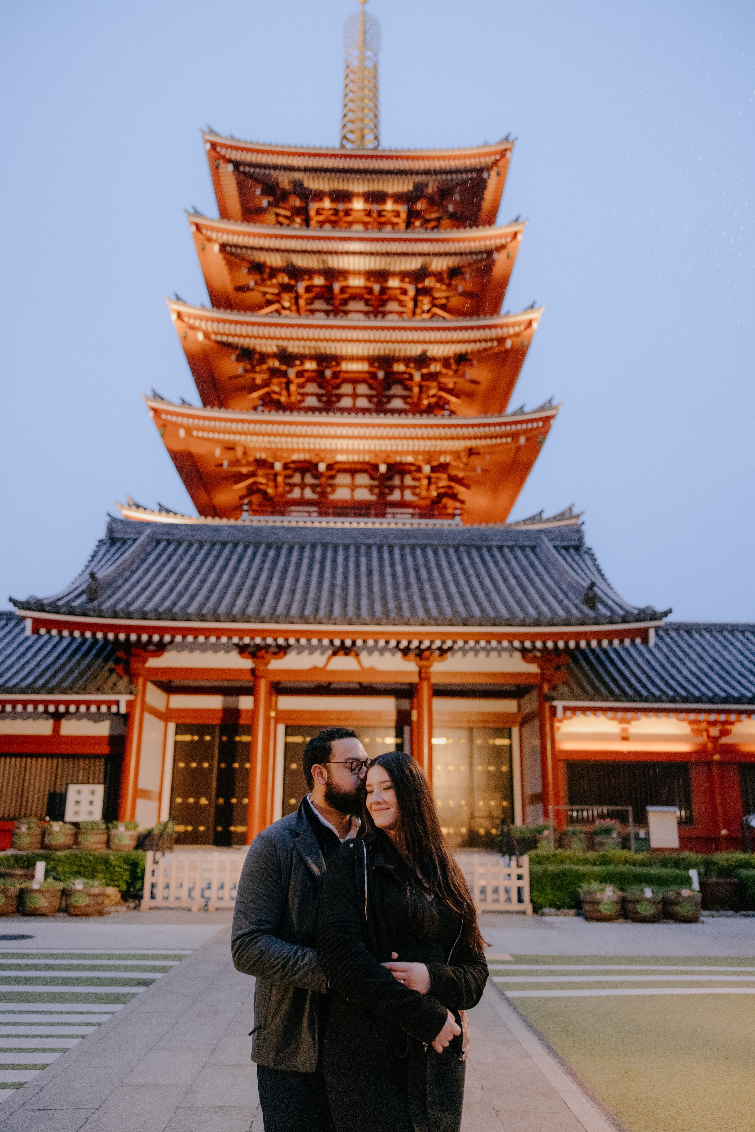 Two people in dark winter clothing stand in front of illuminated Sensoji Temple's five-tiered pagoda at dusk in Tokyo.