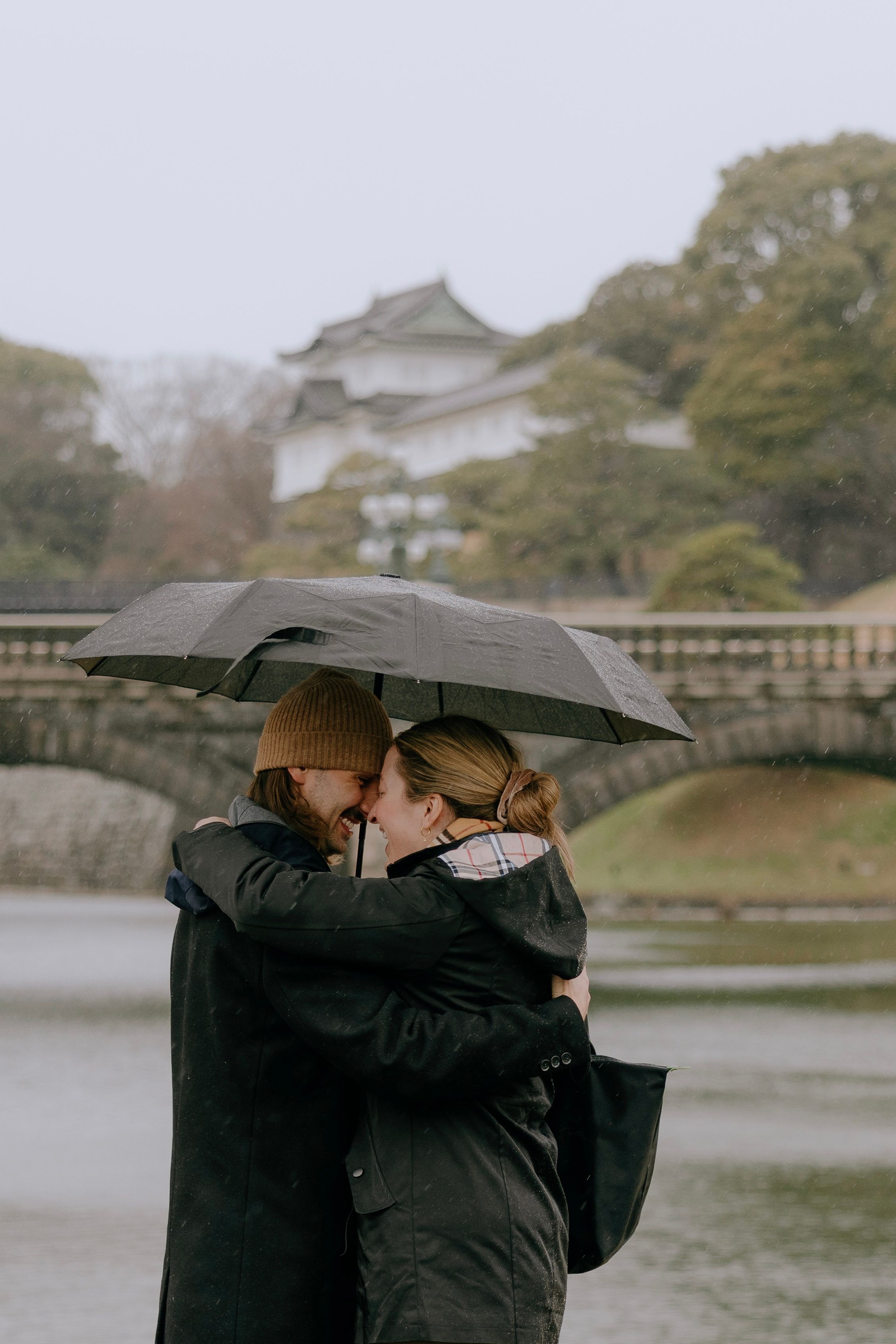 Two people share an intimate moment under an umbrella at a Japanese castle garden on a misty day.