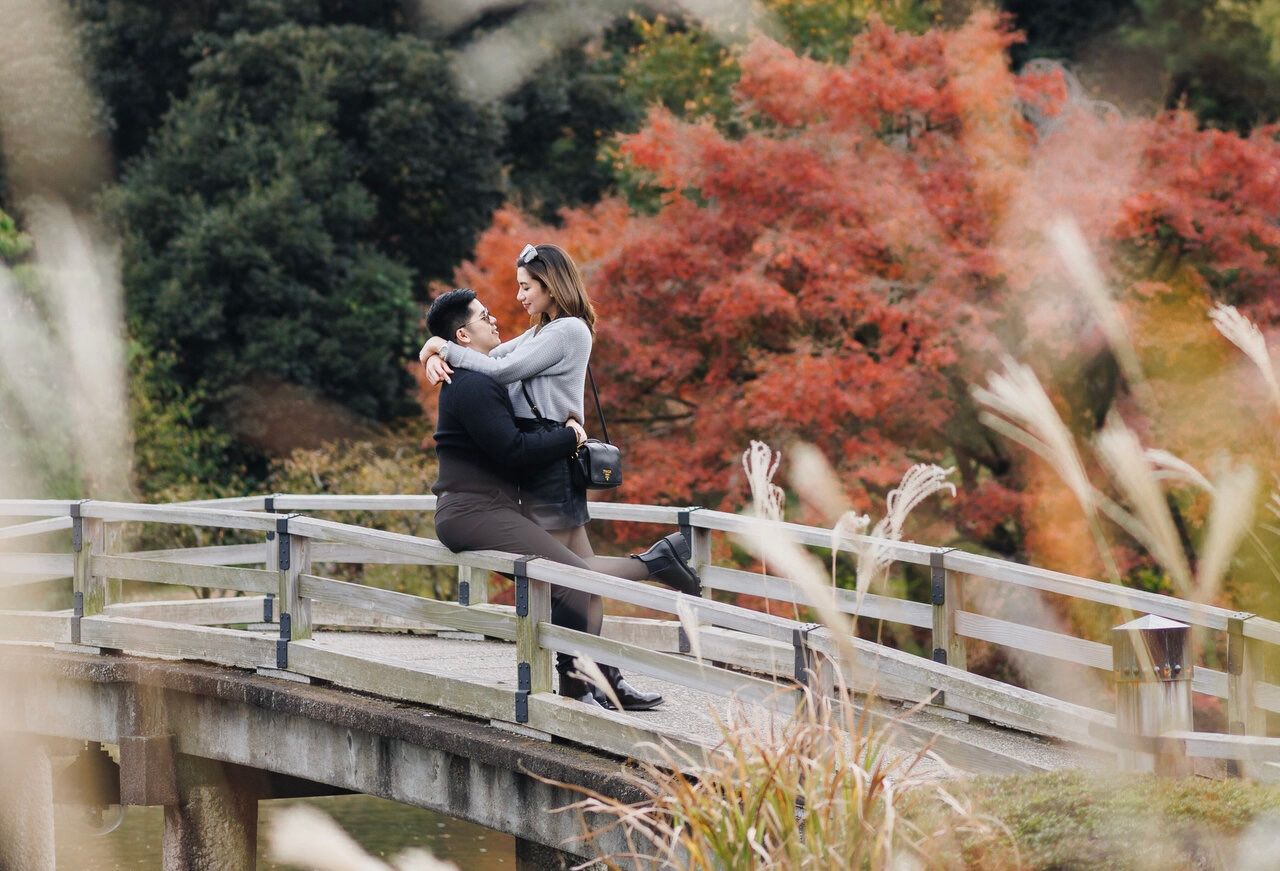 A couple shares a tender moment on a wooden bridge with vibrant red autumn leaves.