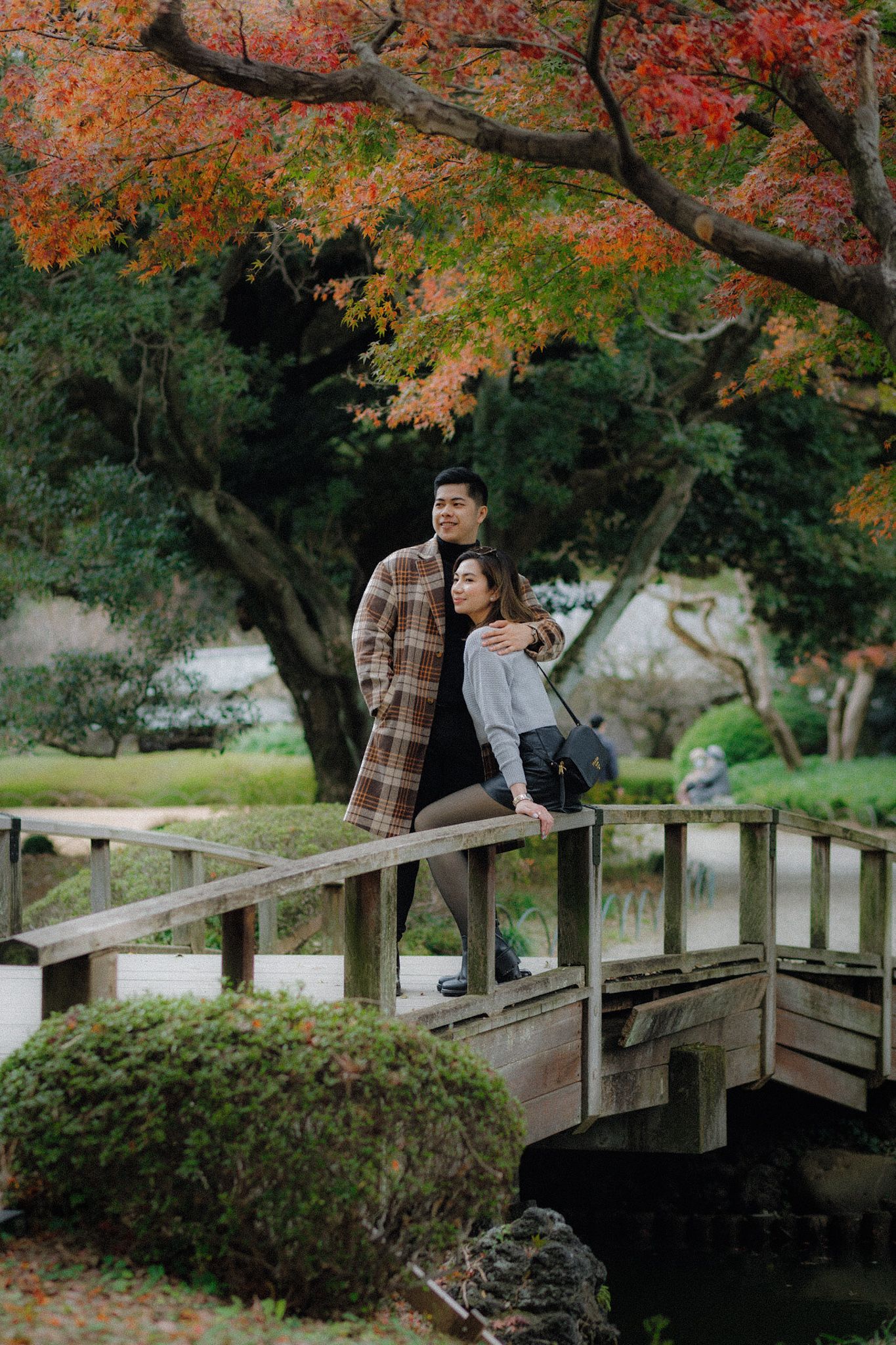 A couple stands on a wooden bridge under beautiful autumn maple trees in a Japanese garden.
