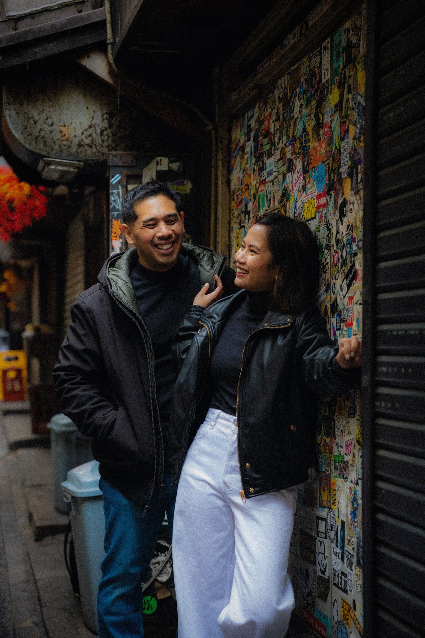 A couple laughs together against a graffiti-covered wall in an urban alleyway.