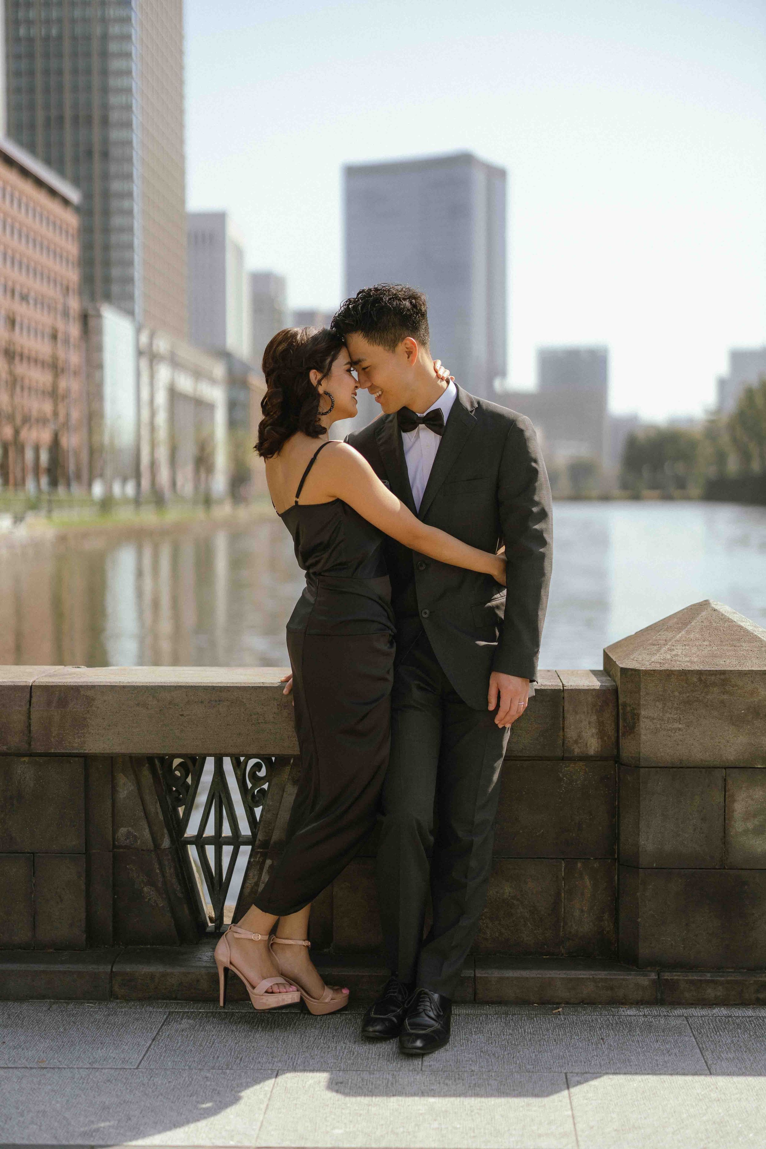A romantic couple in formal wear embraces against an urban cityscape backdrop.