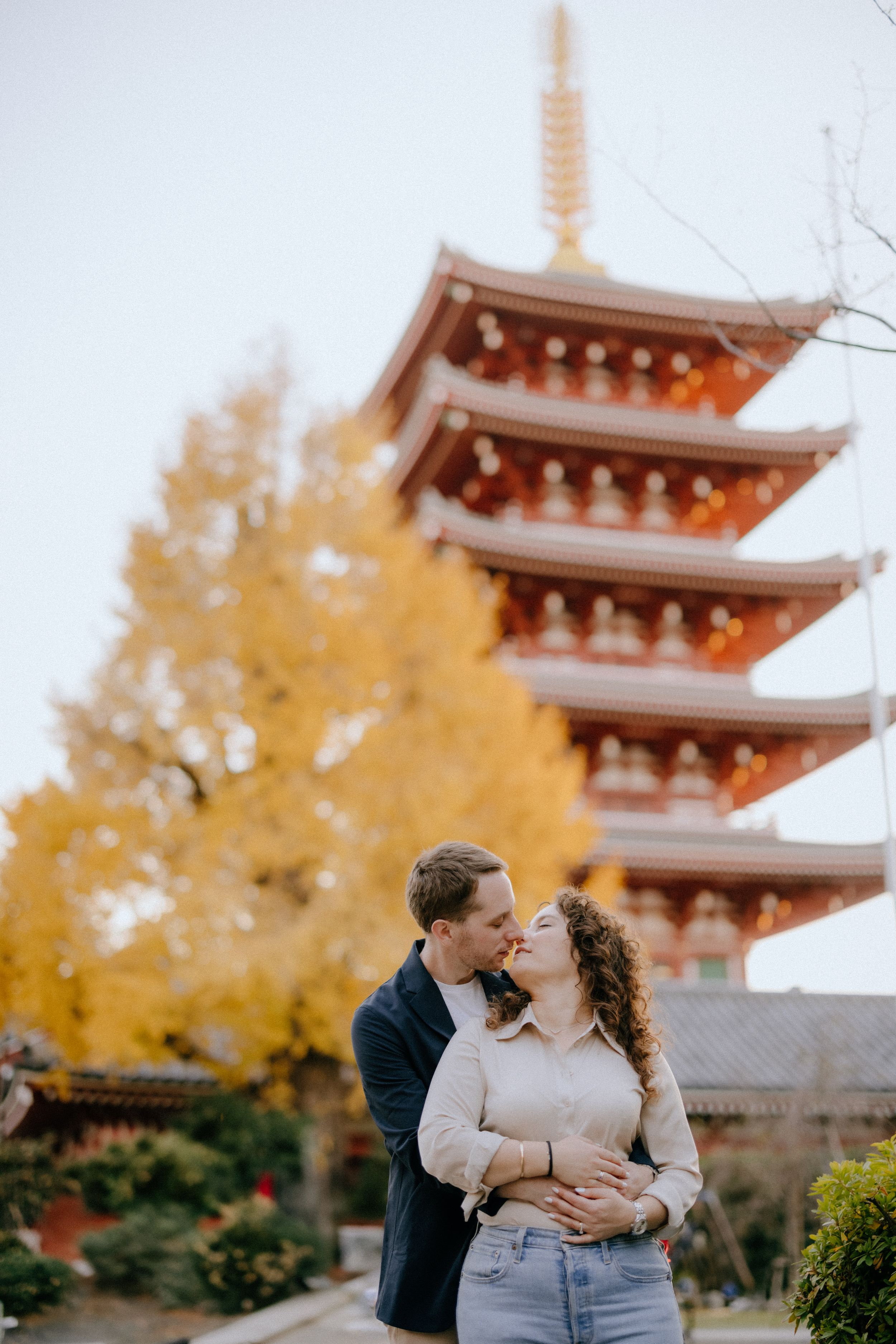 A couple embraces in front of a traditional five-story Japanese pagoda with autumn foliage in the background.