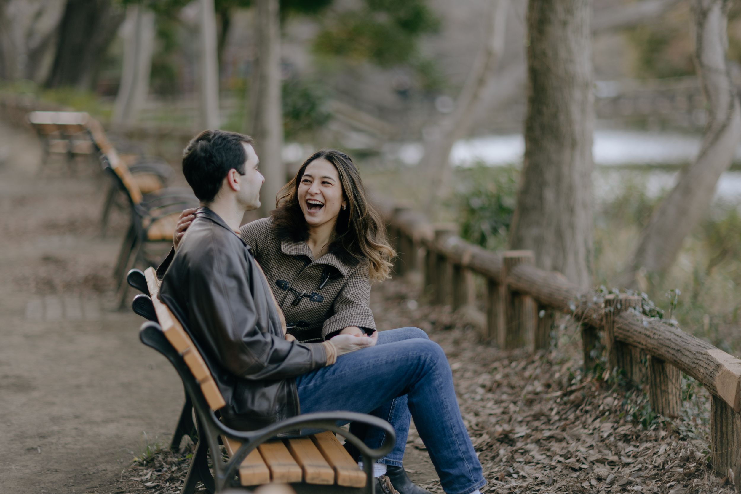 A laughing couple shares a joyful moment while sitting on a wooden bench along a wooded path.