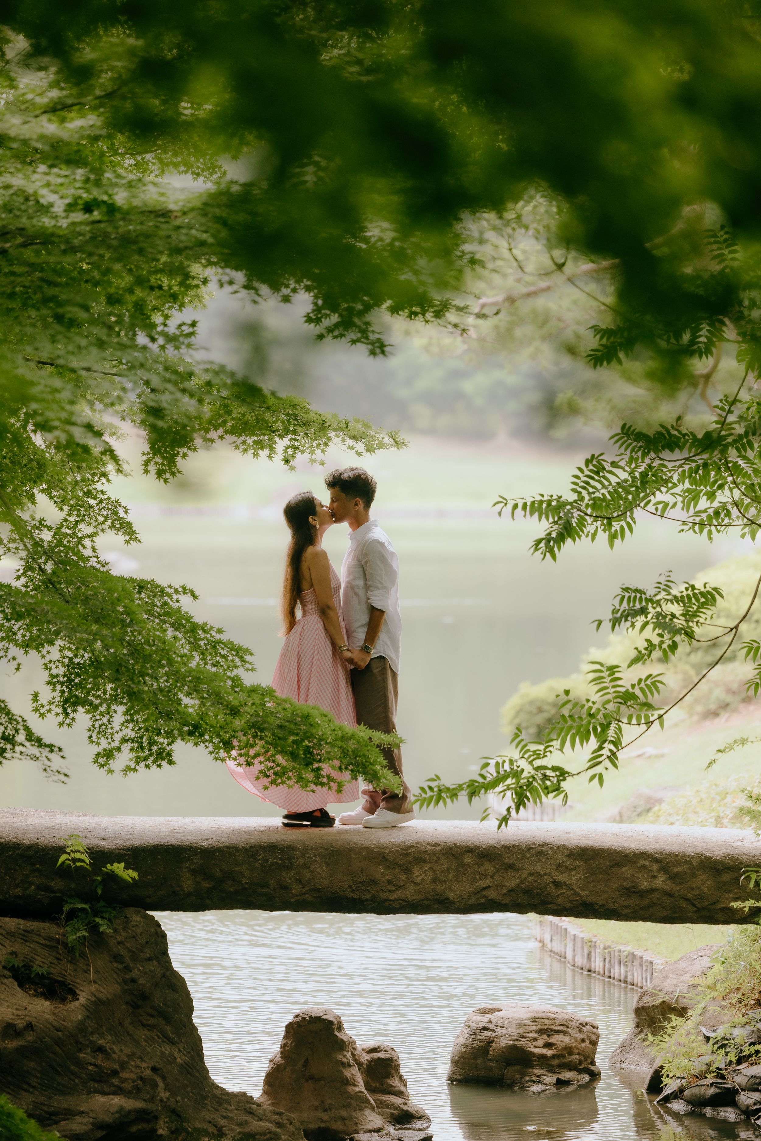 A couple shares a romantic moment on a stone bridge framed by overhanging green tree branches above a serene lake.