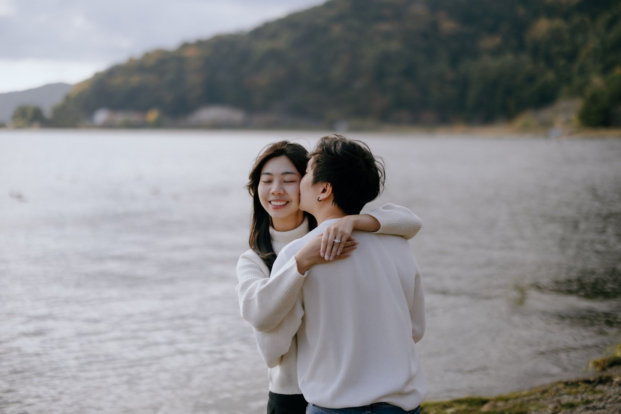 Couple in white sweaters embrace while standing on a serene lakeshore with hills in the background.