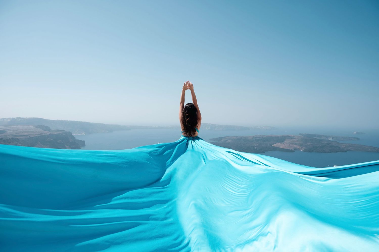 Person holding flowing turquoise fabric against Mediterranean coastal landscape.