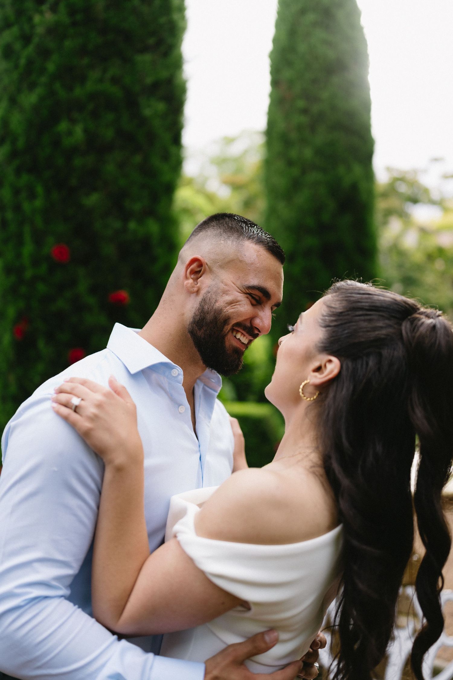 A couple embraces and laughs together in front of tall cypress trees in a romantic garden setting.