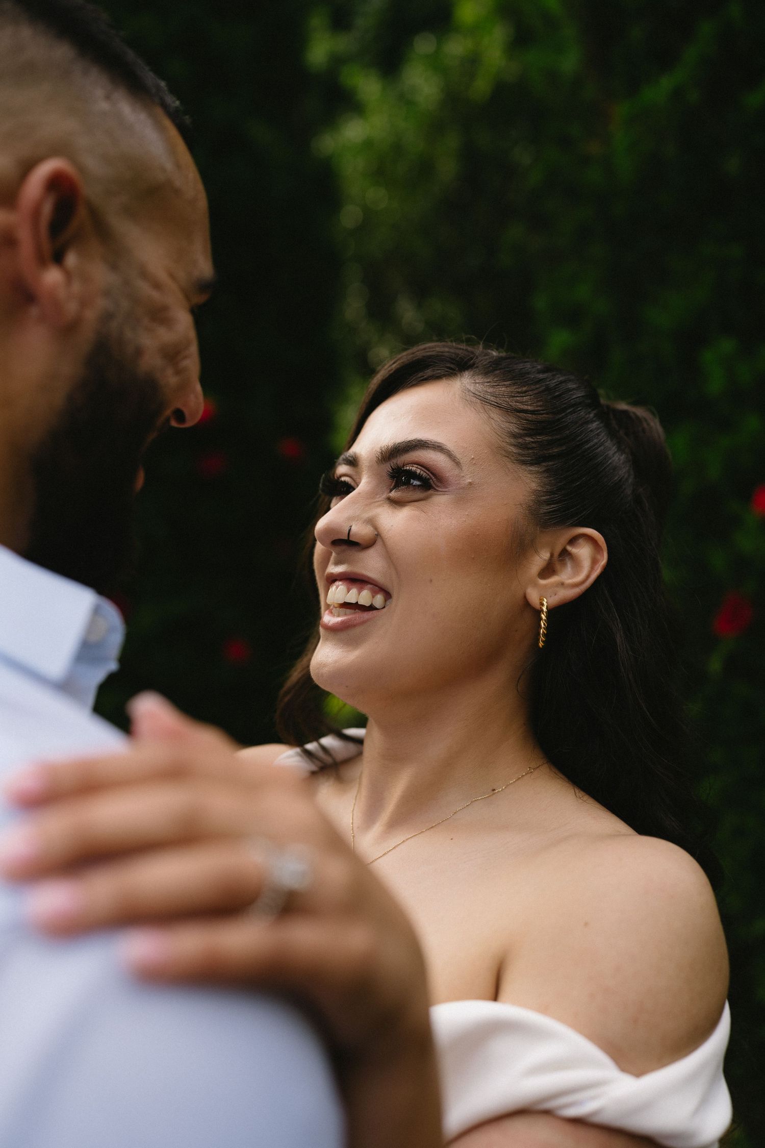 A couple shares an intimate moment in a garden setting, with greenery and red flowers in the background.