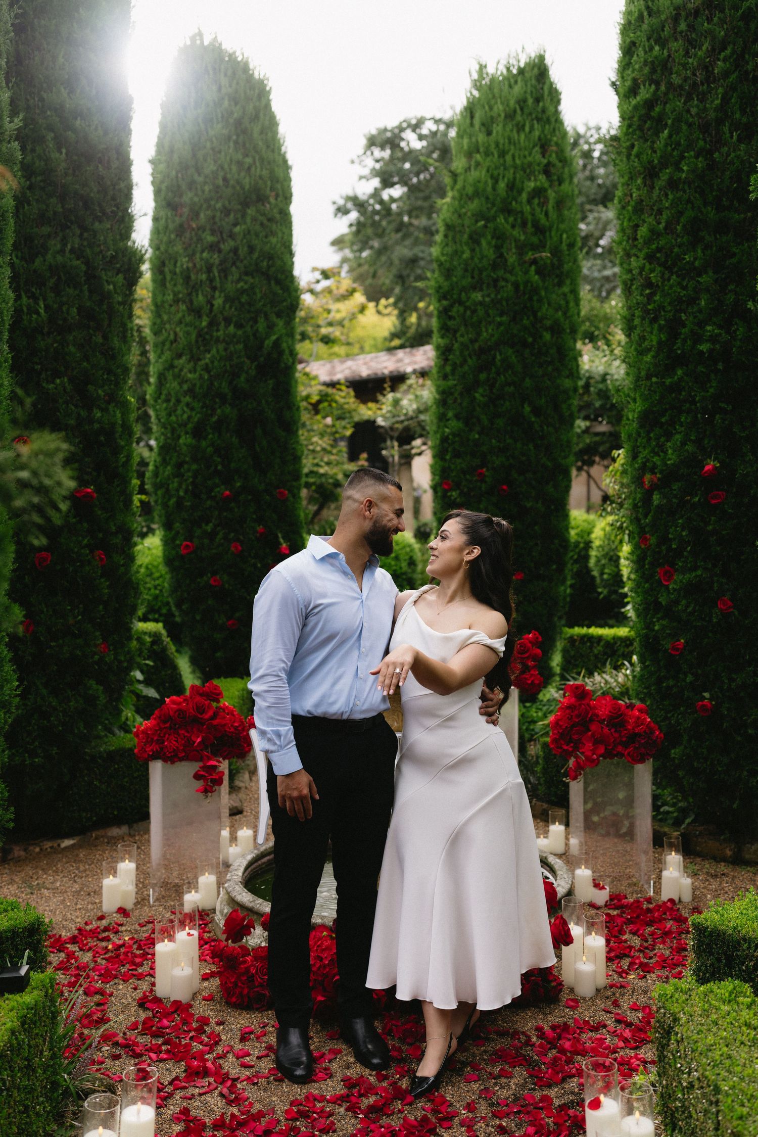 A romantic couple stands on rose petals surrounded by tall cypress trees and candles in a garden.