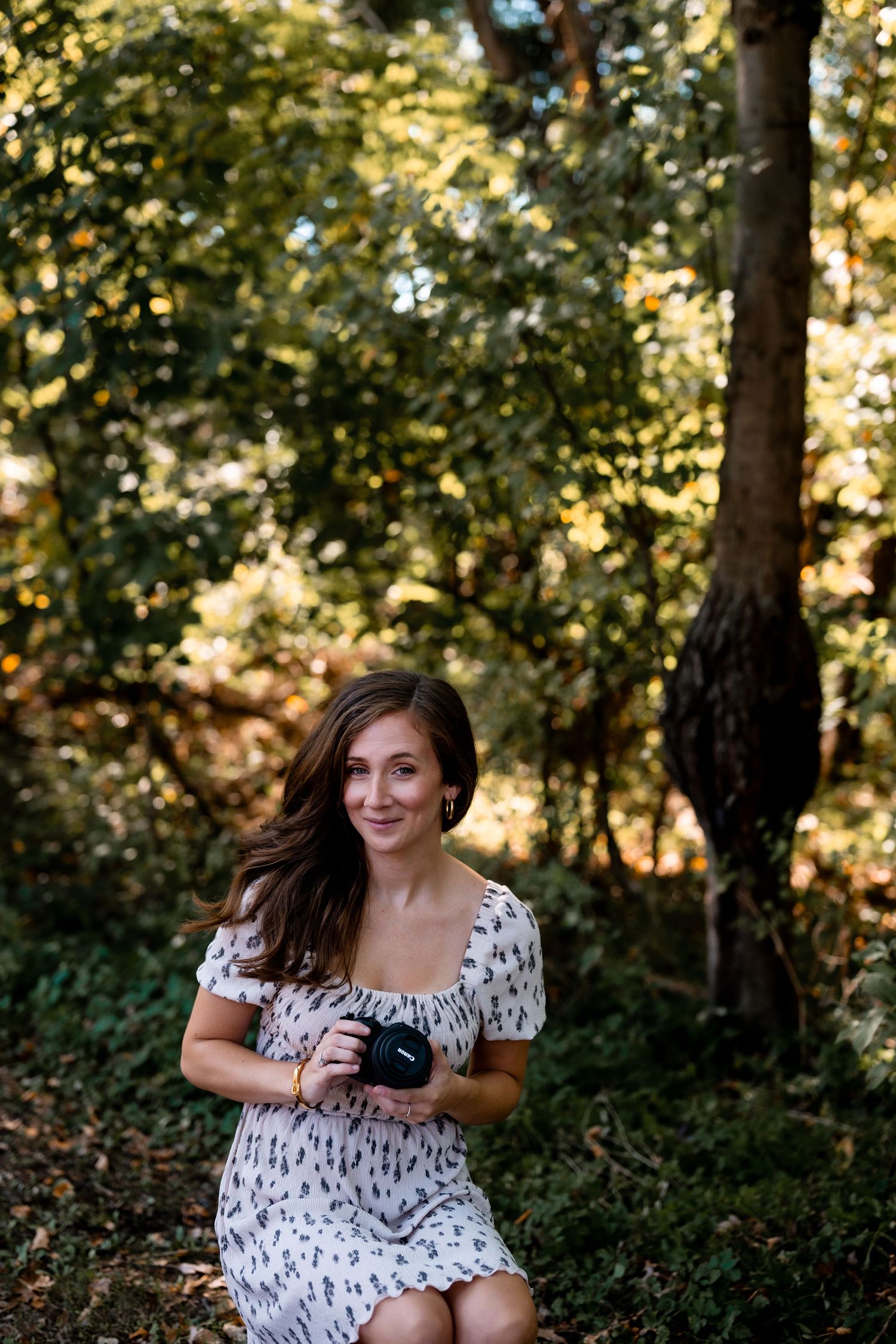 Woman in floral dress sitting in sunlit forest setting.
