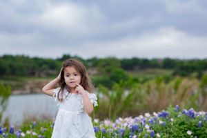 Young girl in white dress stands among bluebonnets with a lake and green foliage in the background. Bluebonnet Mini Session Austin TX Brushy