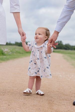 Small child holds hands with adults while walking on dirt path wearing white patterned dress and sandals. bluebonnet Mini session Cedar Park
