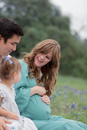 Expecting parents and young child share a tender moment among blooming bluebonnets in an outdoor field. Bluebonnet Mini Session Austin TX