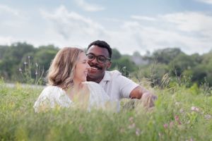 couple sits together in a sunny meadow filled with tall grass and wildflowers on a spring day. Couples Bluebonnet Mini Session Cedar Park TX