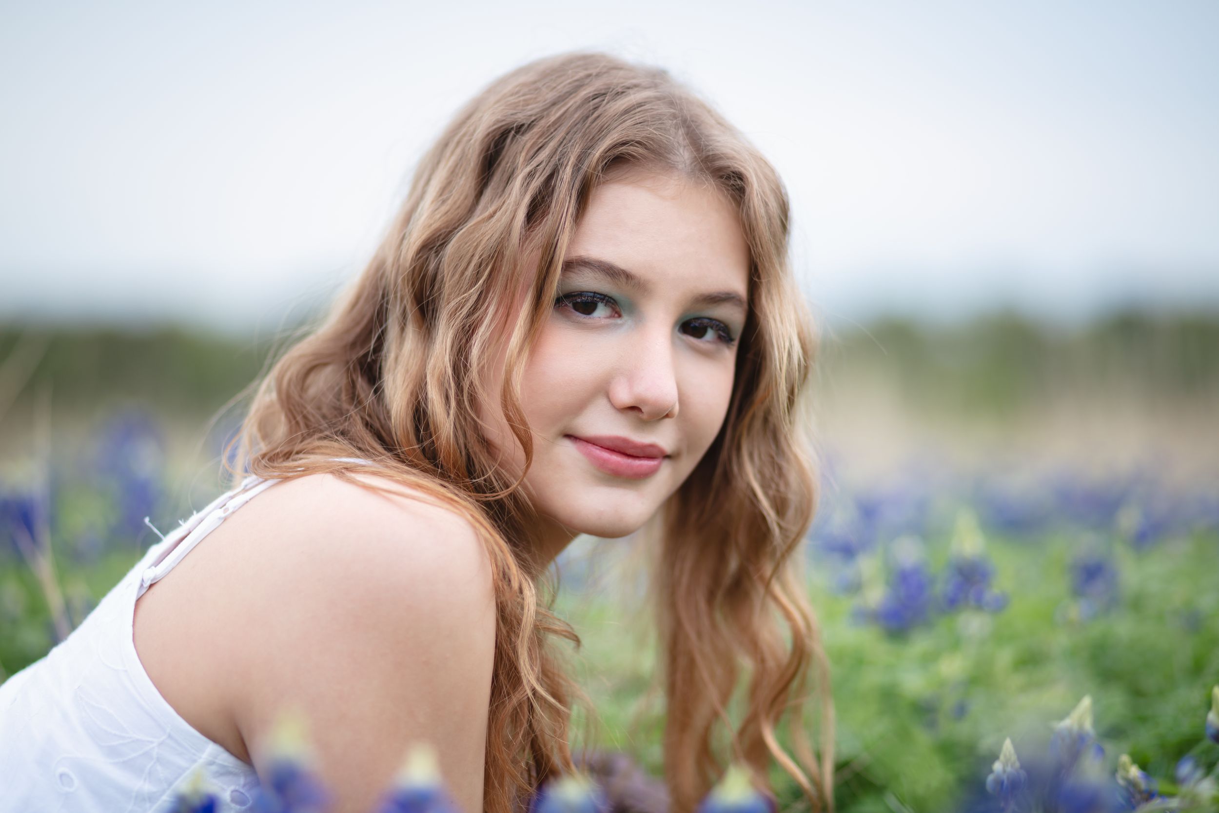 A person with wavy hair poses in a white top among blooming bluebonnet wildflowers on a sunny day. Brushy Creek Lake Park Cedar Park Mini