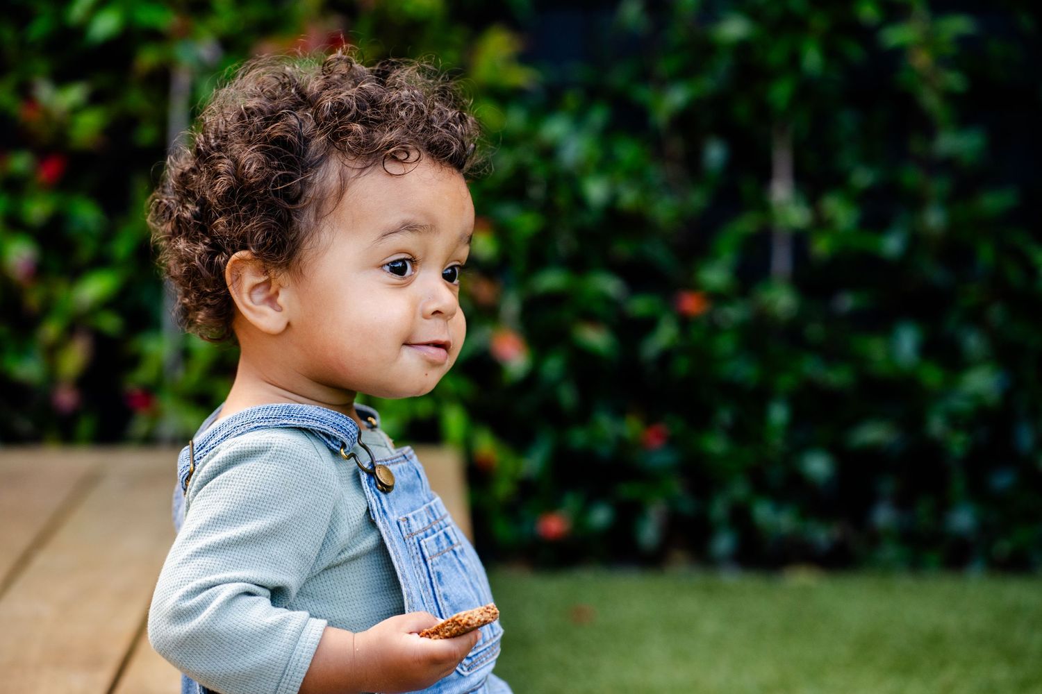 Small child in denim overalls smiling outdoors against green foliage.