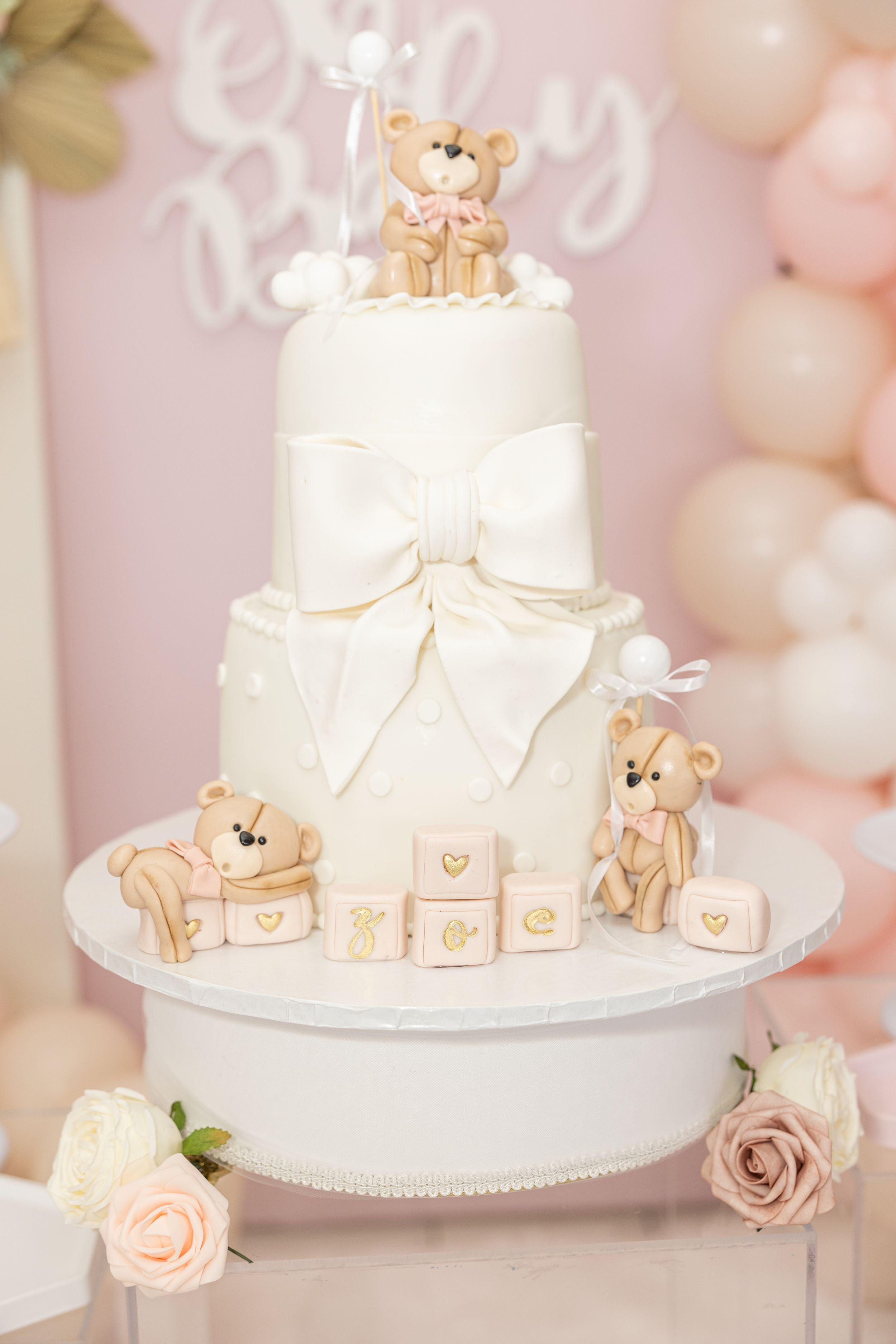 Elegant white tiered baby shower cake decorated with teddy bear accents and bows against pink balloon backdrop.