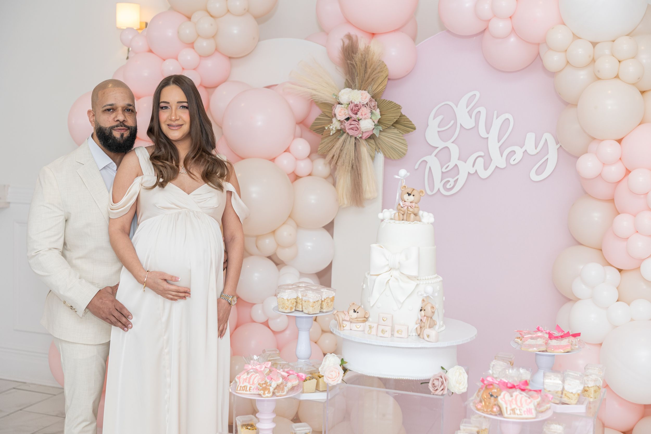 Baby shower celebration setup with white cake, balloon arch and themed decorations in pink and white.