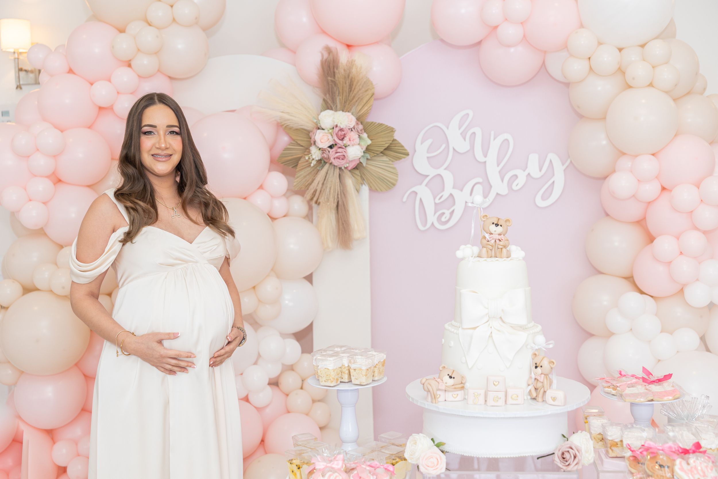 Baby shower setup featuring white tiered cake and pink balloon arch with 'Oh Baby' sign.