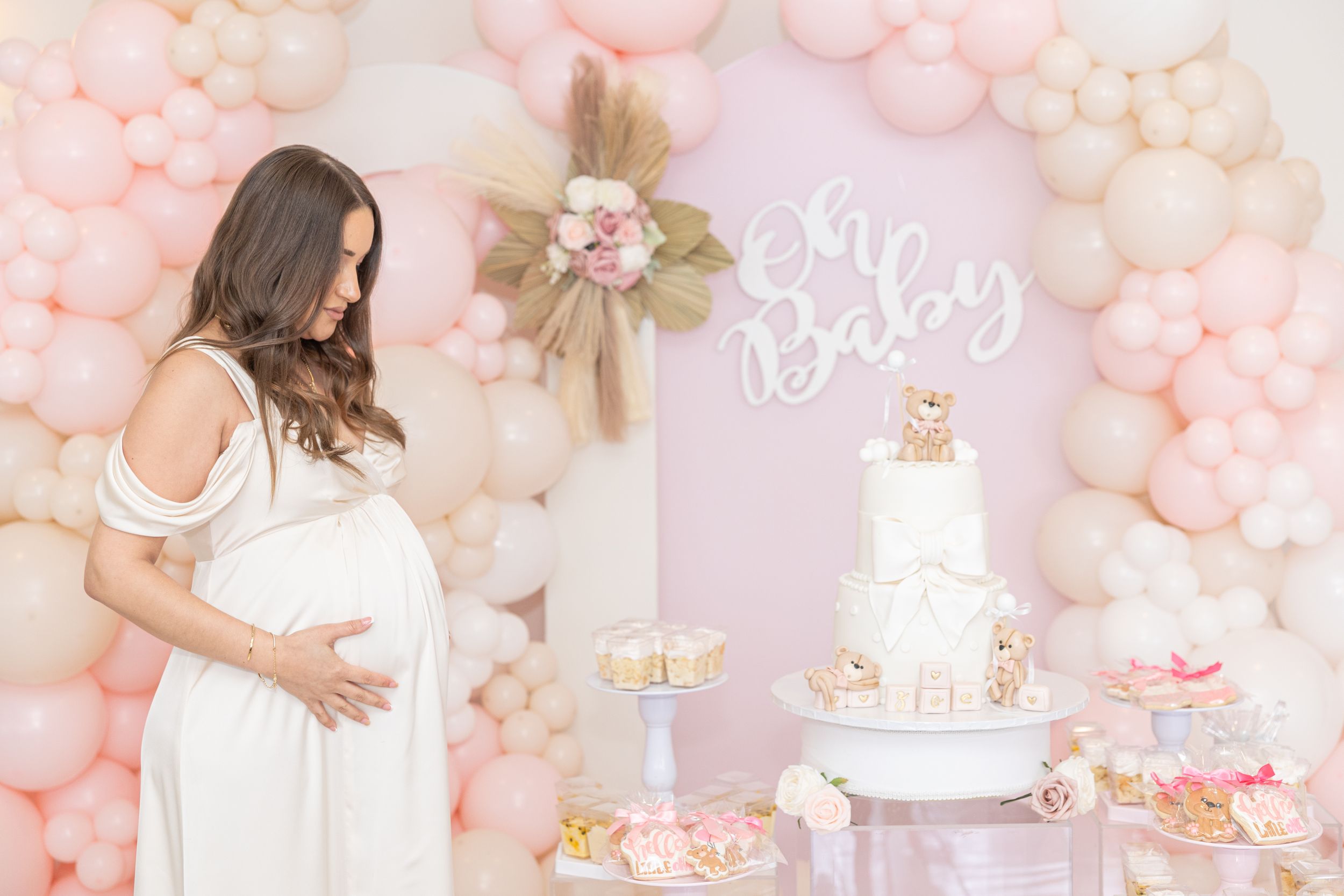 Baby shower setup with elegant white cake, pink balloon arch, and 'Oh Baby' sign in soft pink tones.