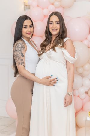 Two women pose together against a pink balloon backdrop at a baby shower celebration.