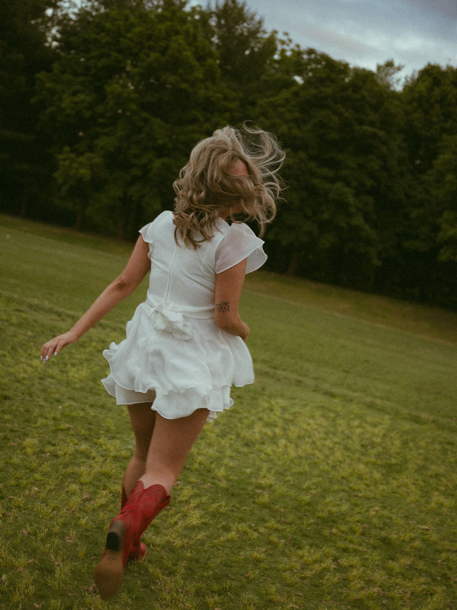 Woman in white ruffled dress and red cowboy boots running through a green field with trees in background.