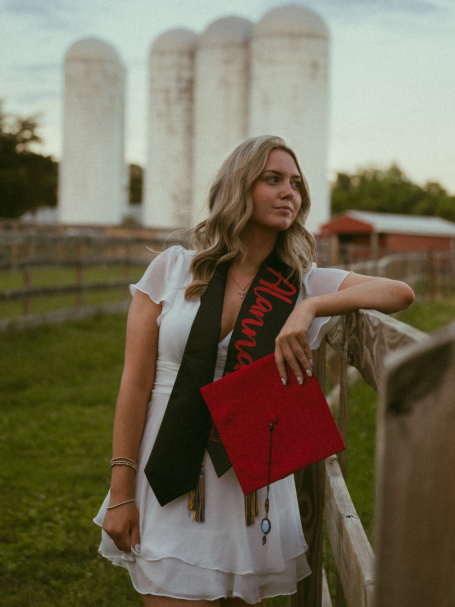 Young woman in white dress holding red graduation cap with black sash, posing near farm fence with silos in background.