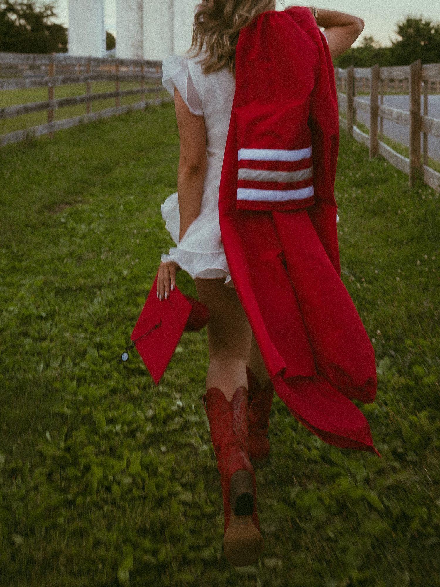 Graduate in red gown and cowboy boots holding cap on green farm, walking away from camera.