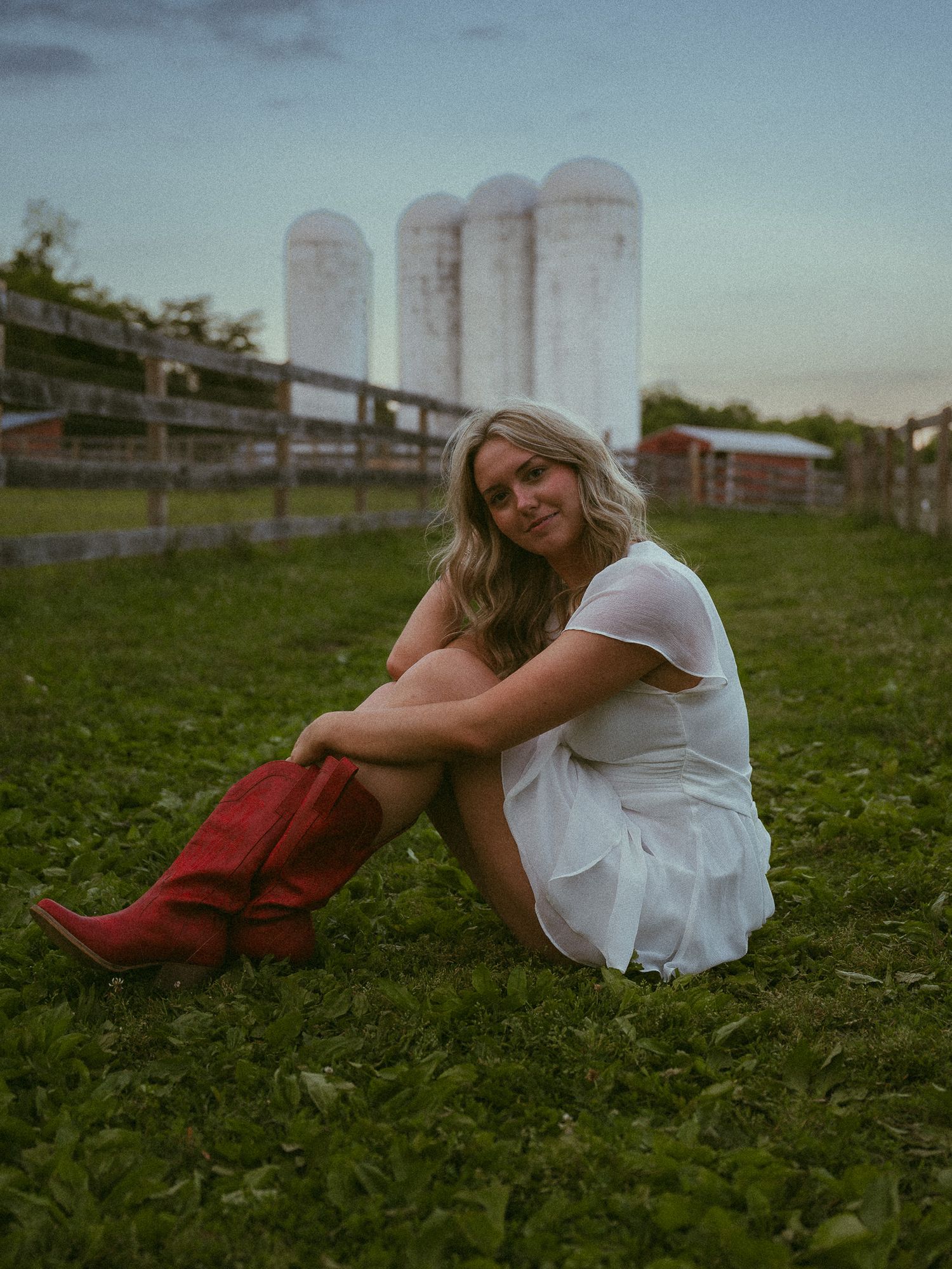 Young woman in white dress and red cowboy boots sitting on green grass with white farm silos in background.