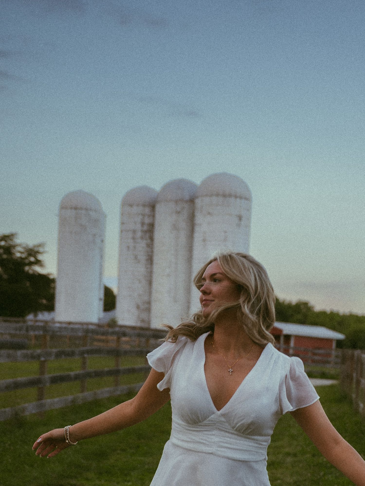 Blonde woman in white wrap dress twirling near white farm silos at dusk on a rural property.