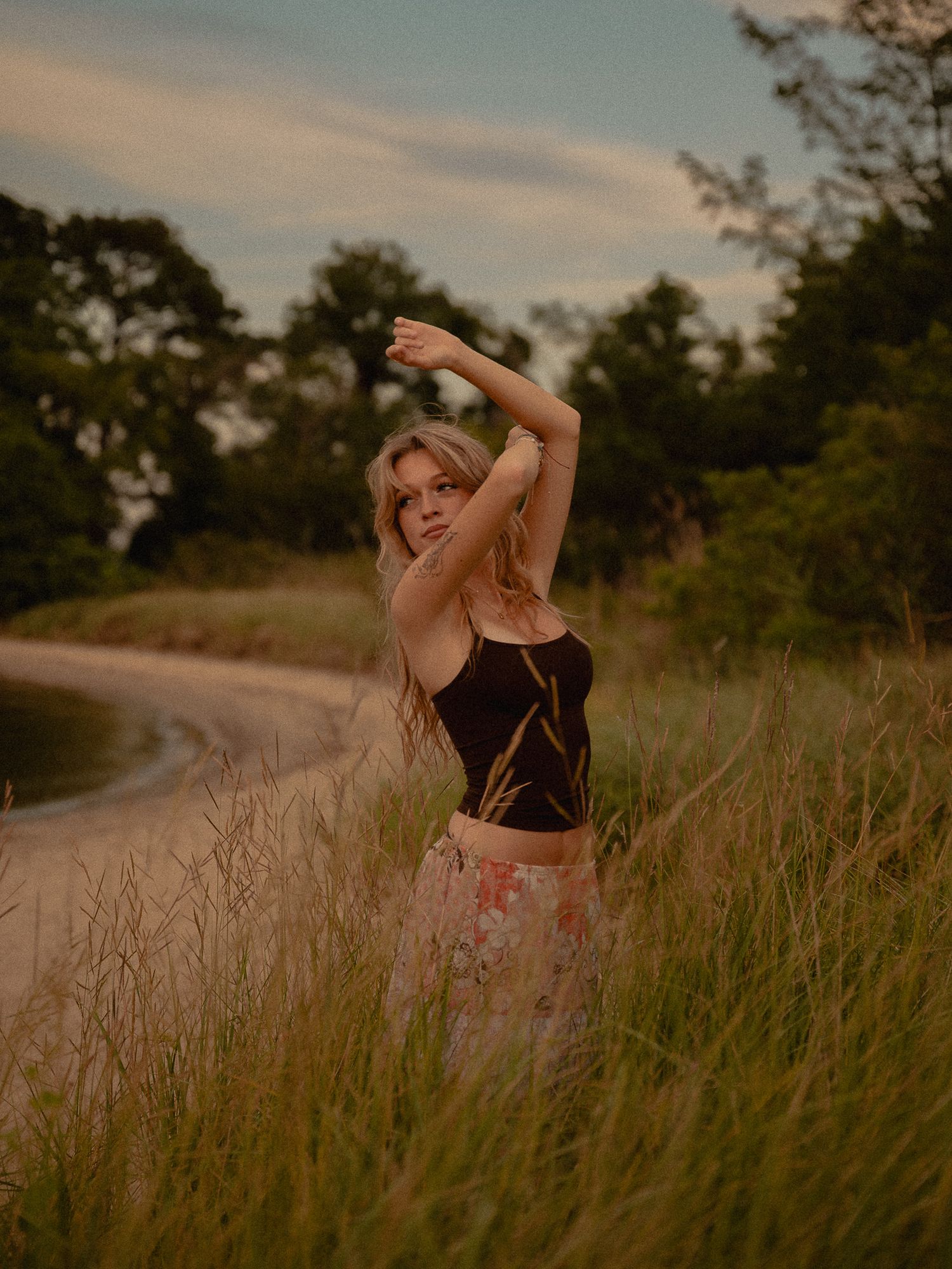 Young blonde woman in black top and floral skirt posing gracefully in tall grass near a sandy path at sunset.