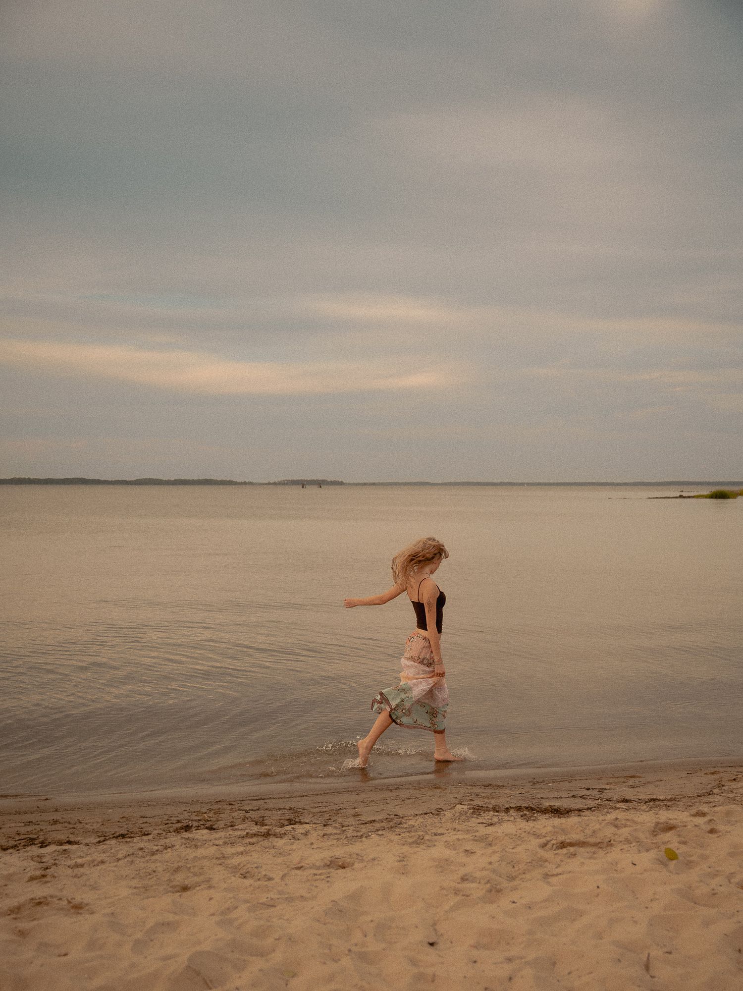 A young blonde girl runs barefoot along a sandy beach shoreline under a cloudy sky.