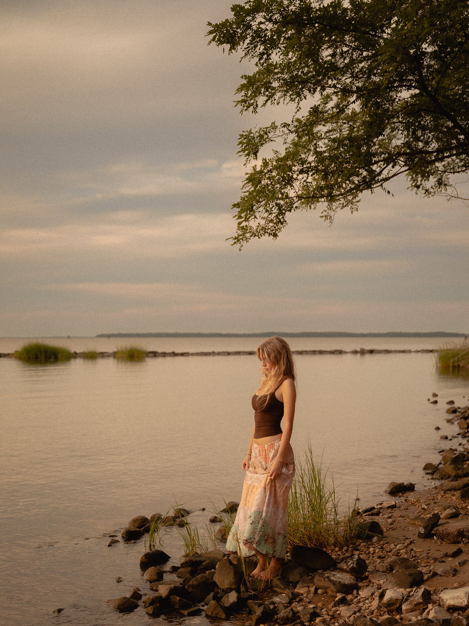 A woman in a floral skirt stands by a calm lake shore at golden hour, with a tree overhead and grassy marshes beyond.