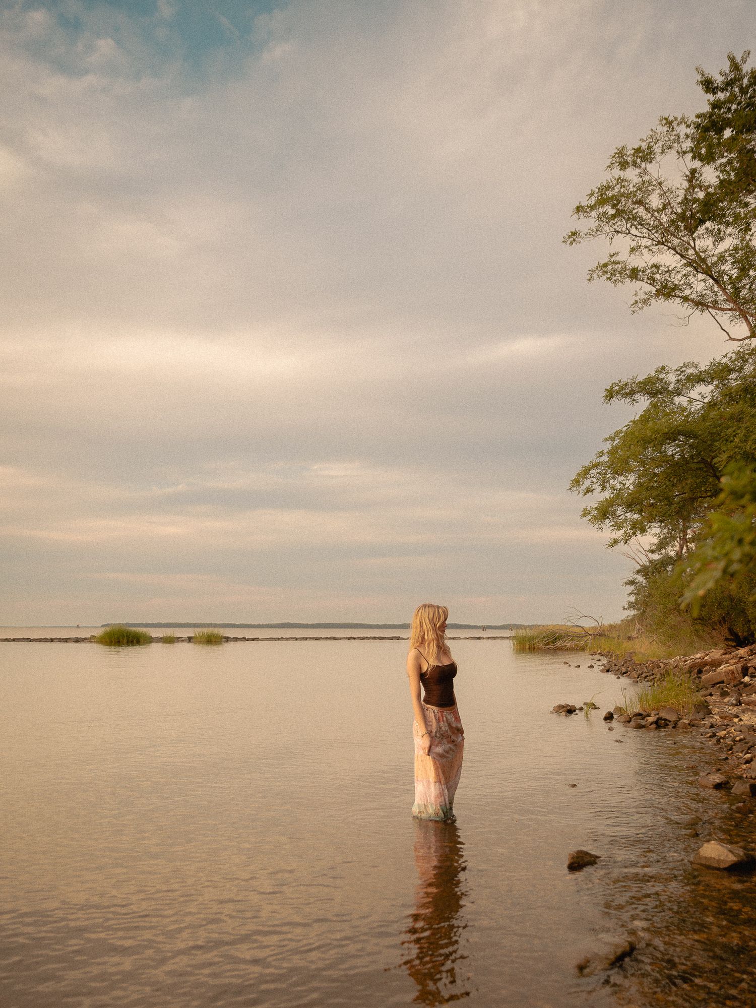 A blonde woman stands waist-deep in calm lake waters near a rocky shore with lush green trees under a cloudy sky.