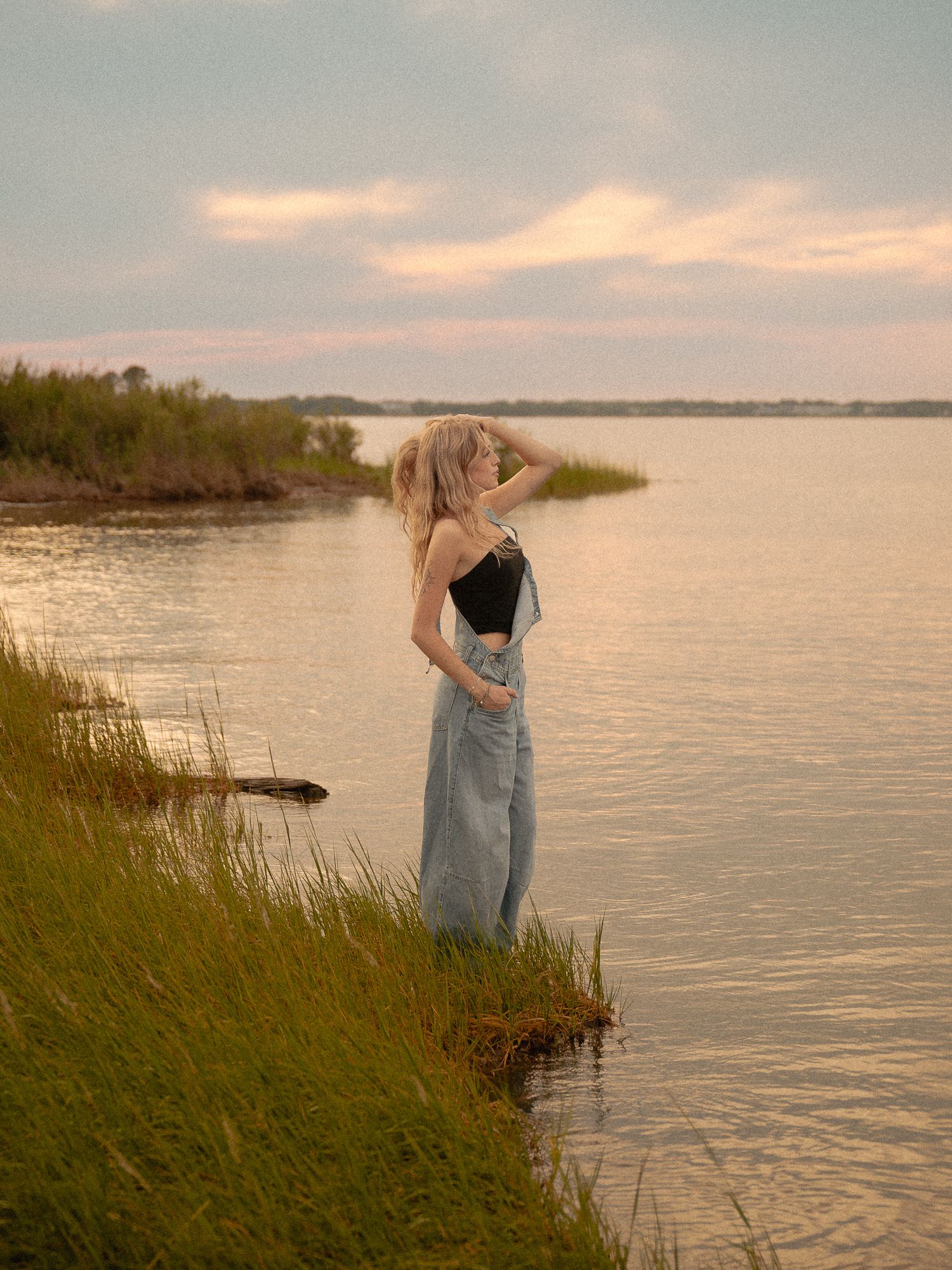 Blonde woman in black top and denim overalls standing by a calm lake at sunset with marsh grass.