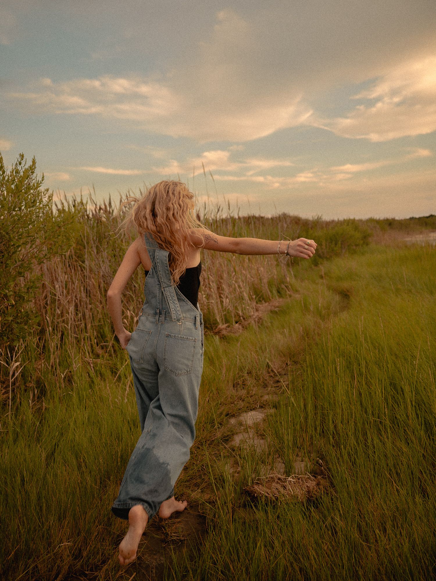 A barefoot blonde woman in denim overalls walks through a lush green field path at golden hour.