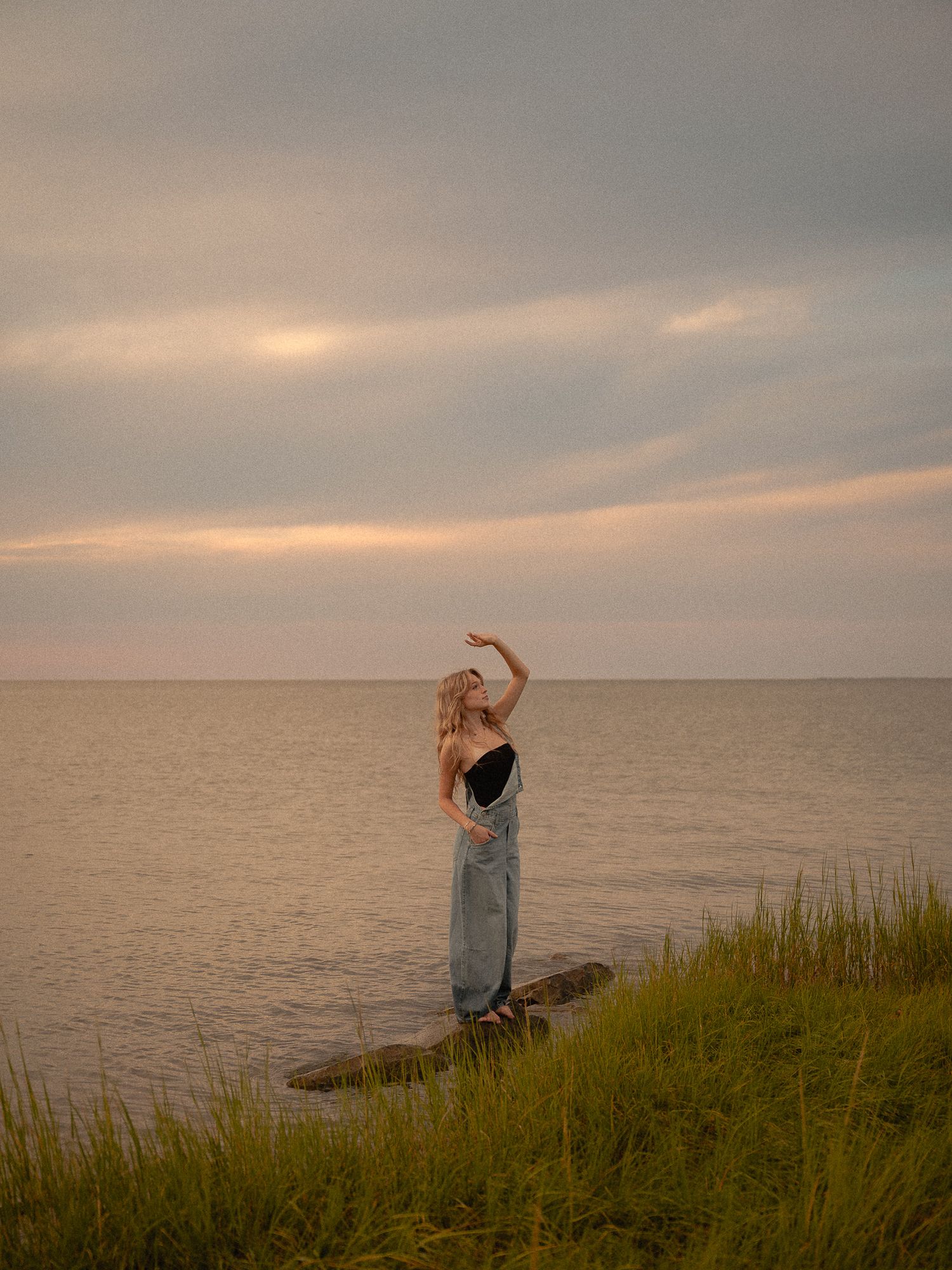 A woman in a black top and blue skirt poses gracefully on rocks by the sea at sunset.