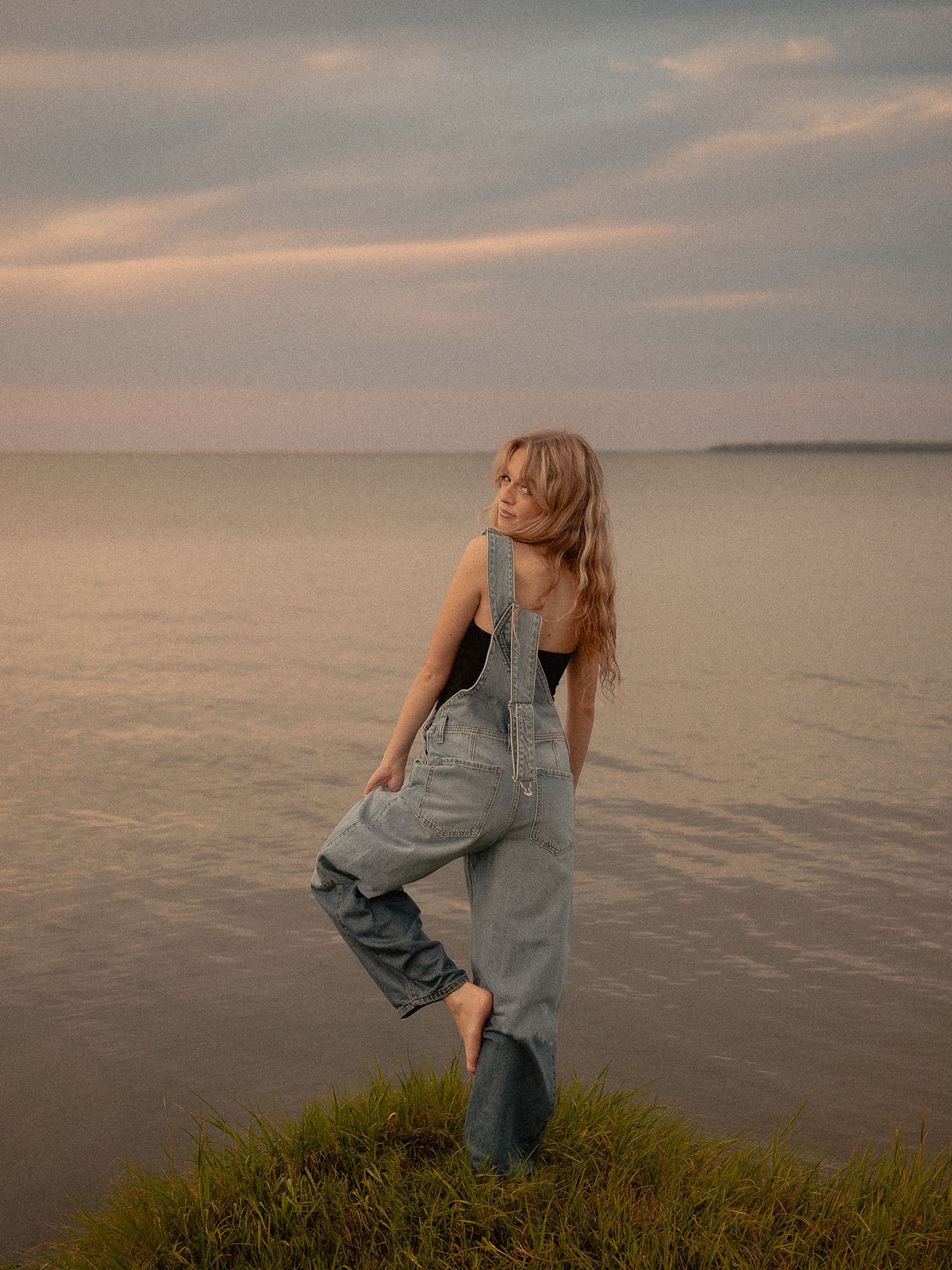 Woman in denim overalls poses on grassy shore at sunset with calm water and cloudy sky in background.
