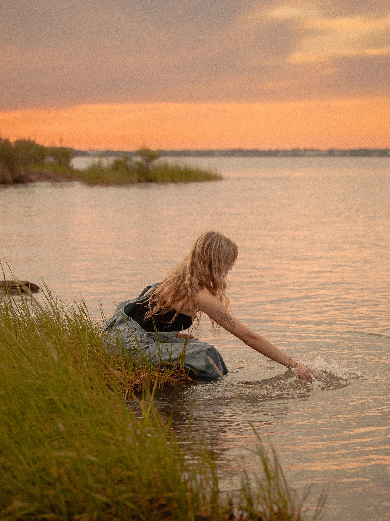 A blonde woman in a dark outfit reaches into calm water at sunset, with golden orange skies reflecting on a serene lake.