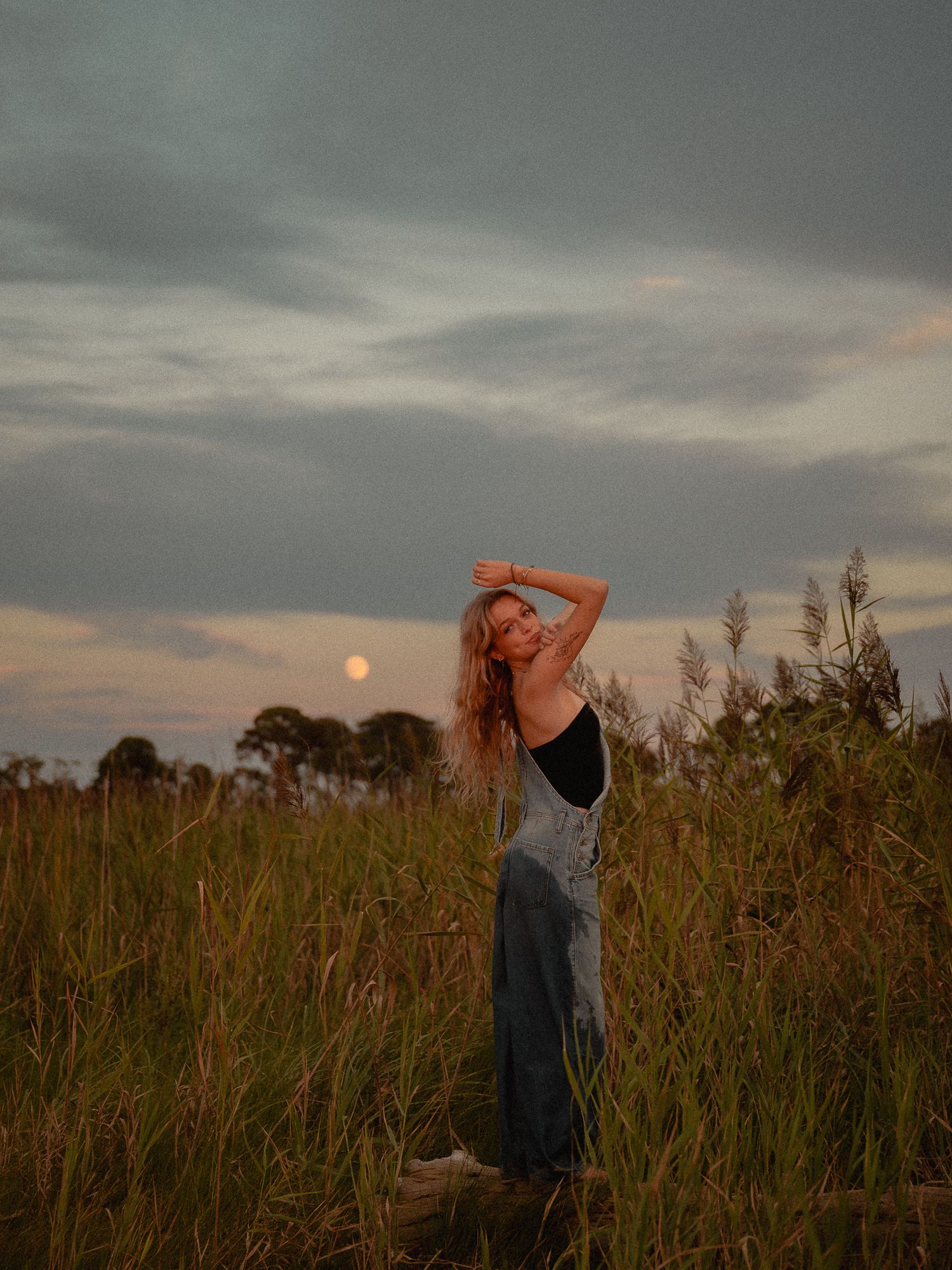 Woman in denim skirt poses in tall grass field at golden hour with full moon rising behind her.
