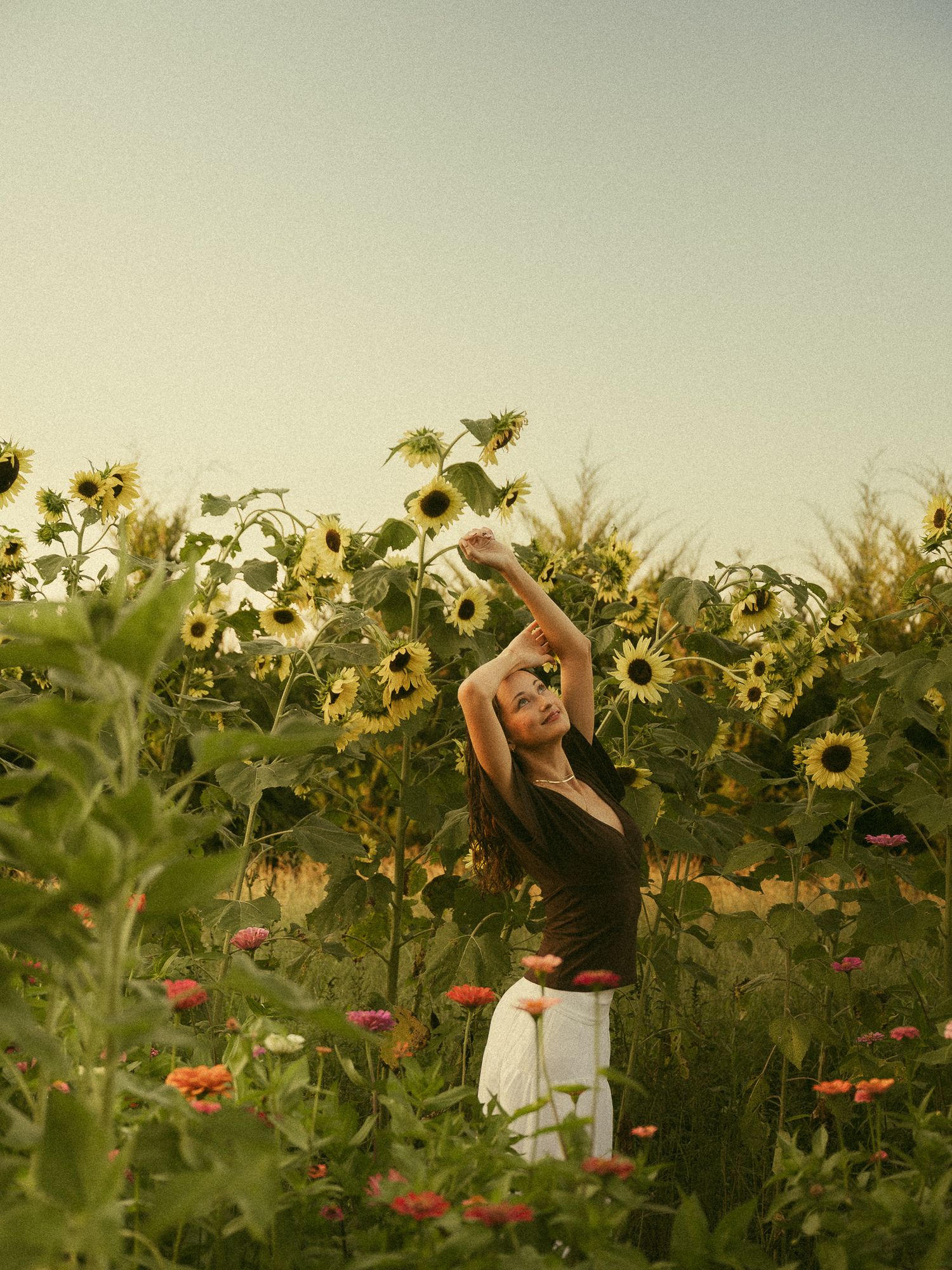 A woman in a black top and white skirt reaches up among tall sunflowers in a lush garden field.