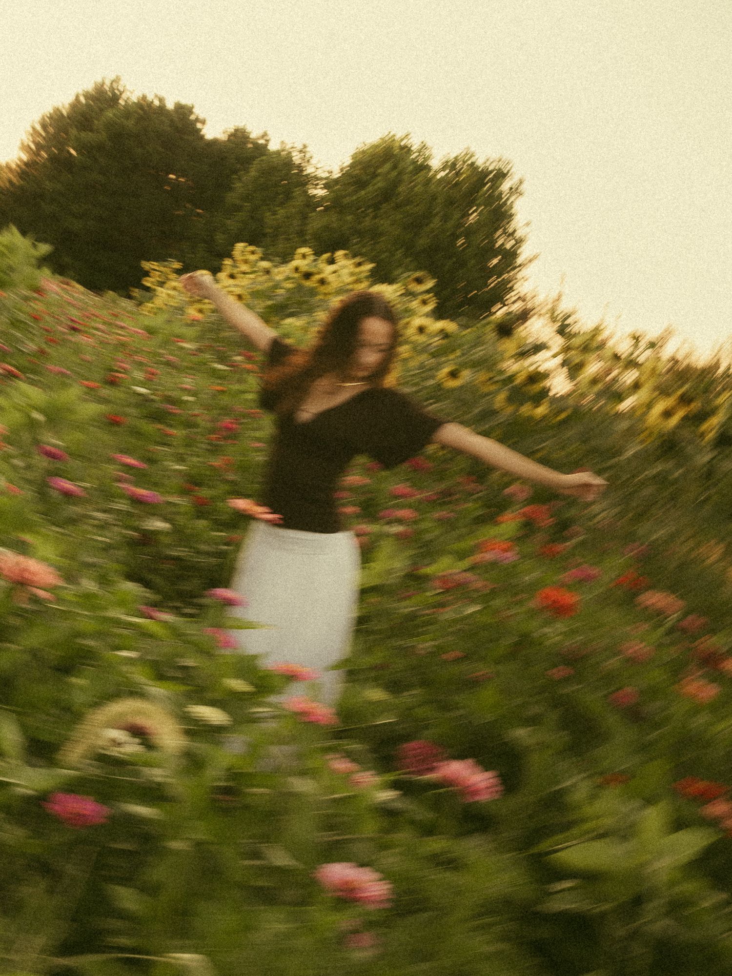 A woman in a black top and white skirt dances joyfully among colorful flowers and sunflowers in a lush garden.