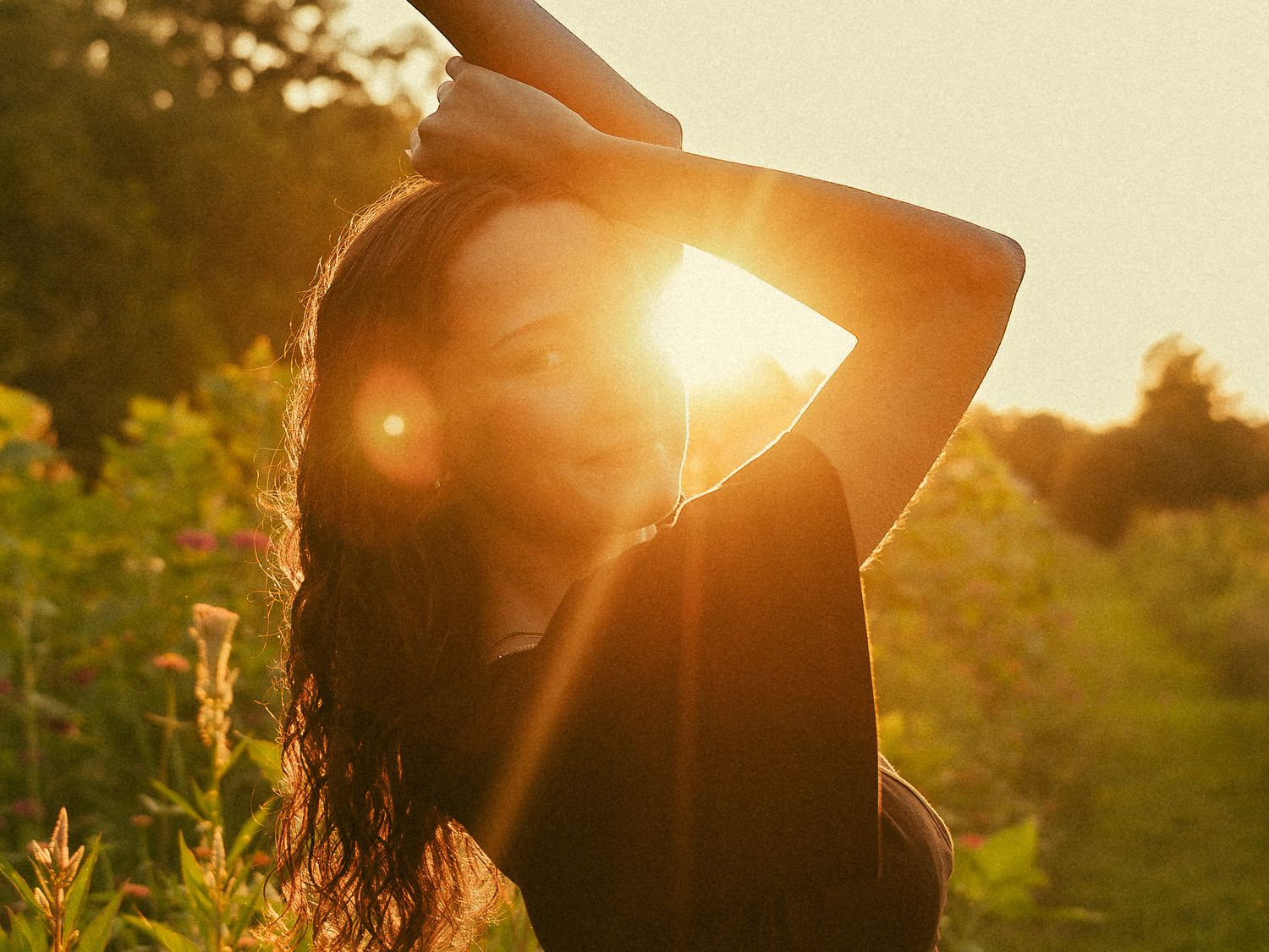 Young woman with long dark hair poses in a sunlit field at golden hour with lens flare and lush greenery.