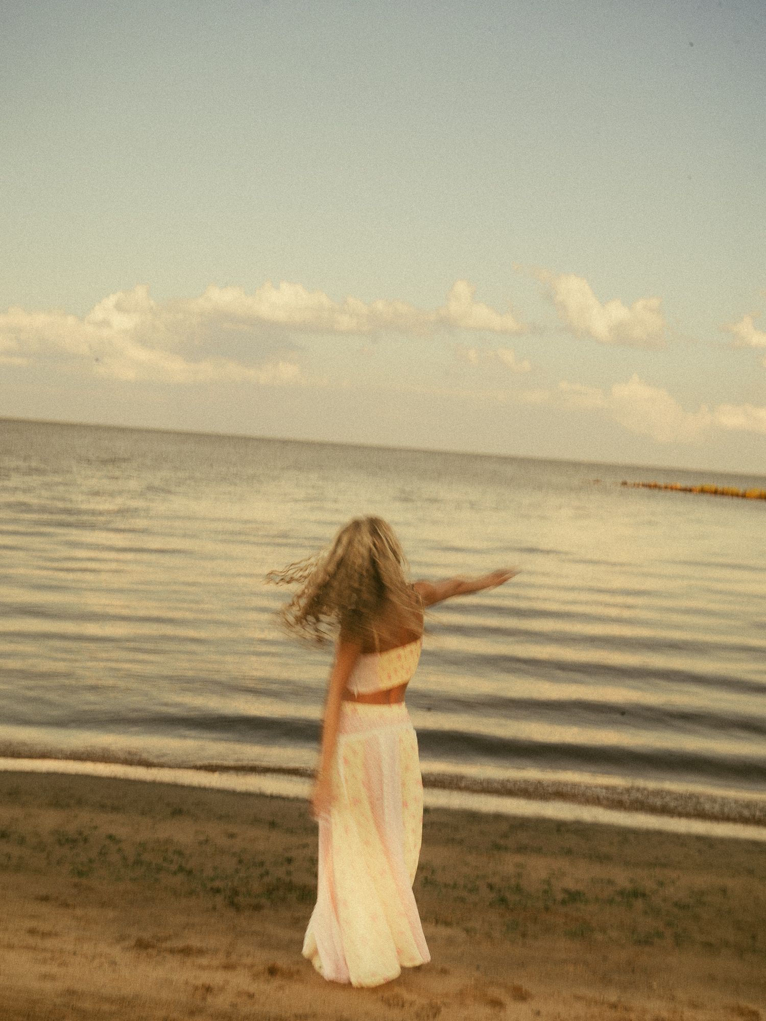 A blonde woman in a white maxi skirt and crop top twirls on a sandy beach with calm ocean waters behind her.